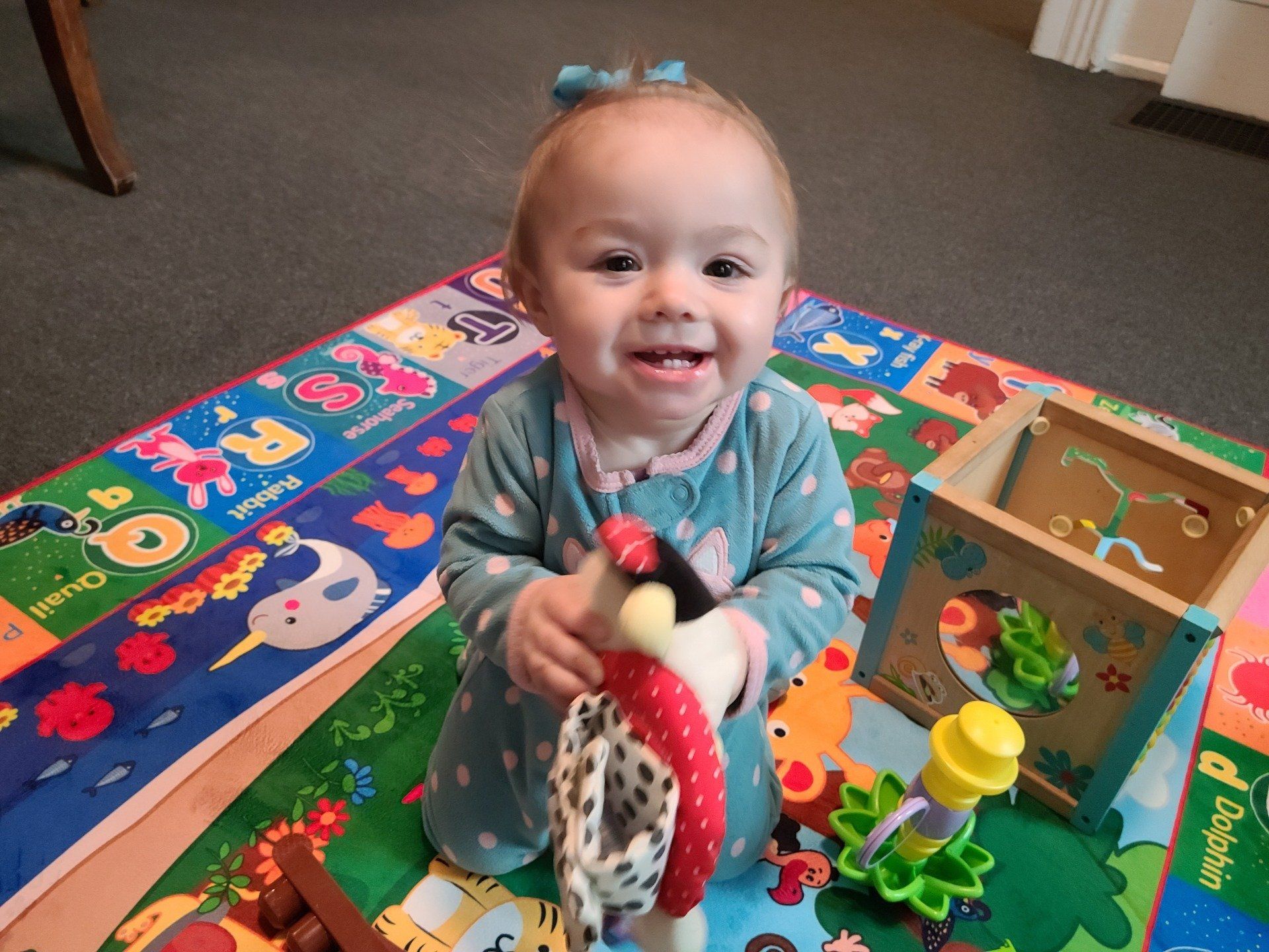 Smiling baby wearing blue, holding toy, sitting on colorful play mat, with toys nearby.