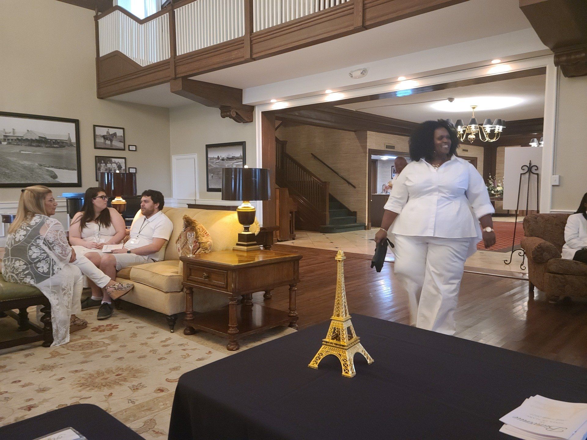 Woman in white suit walks towards camera in a well-lit room with other people seated.
