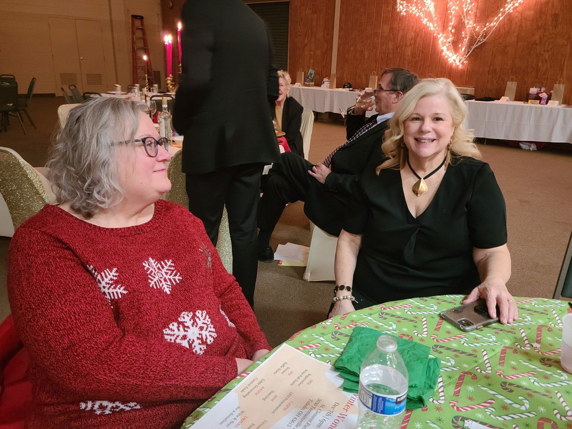 Two women at a festive event, one in a red sweater, the other in black; both smiling at a table.