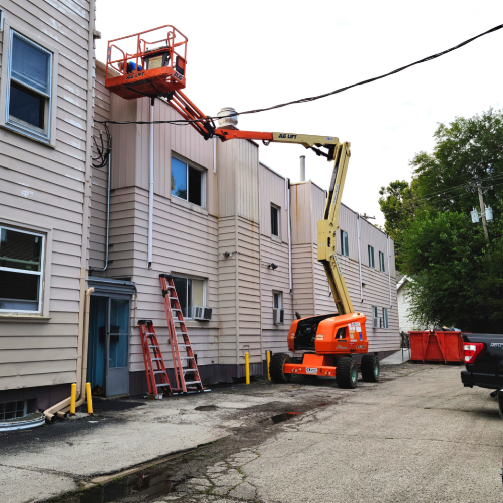 A construction worker in a lift platform working on the side of a light-colored building, next to power lines.