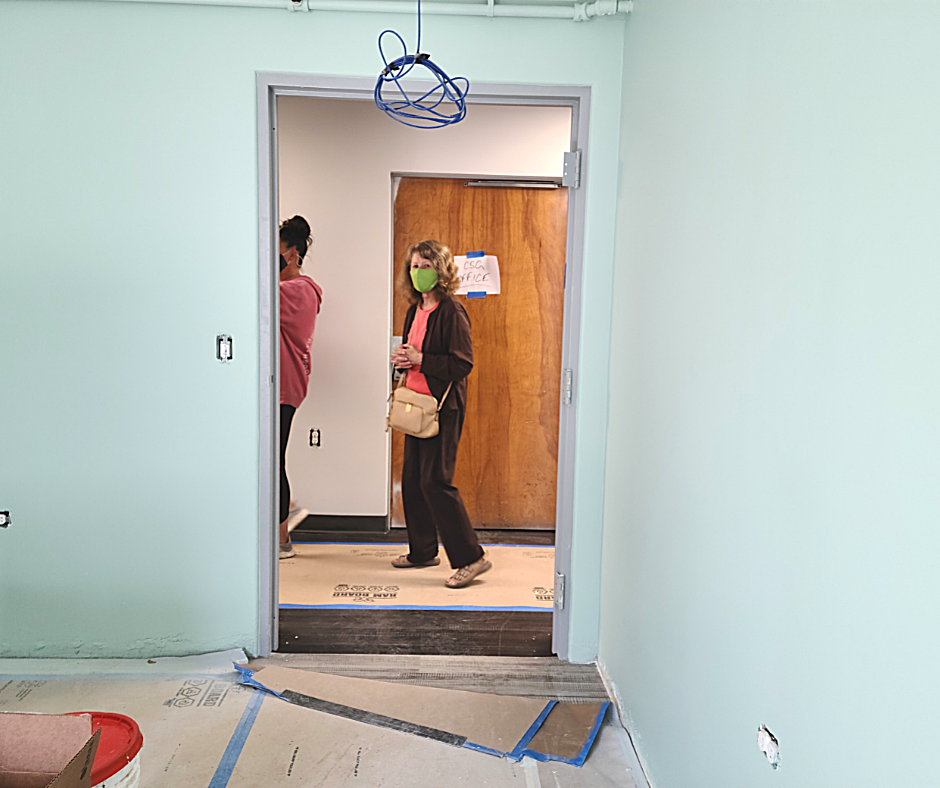 Two women in a doorway; one wears a mask. Light green walls, wooden door, construction floor.