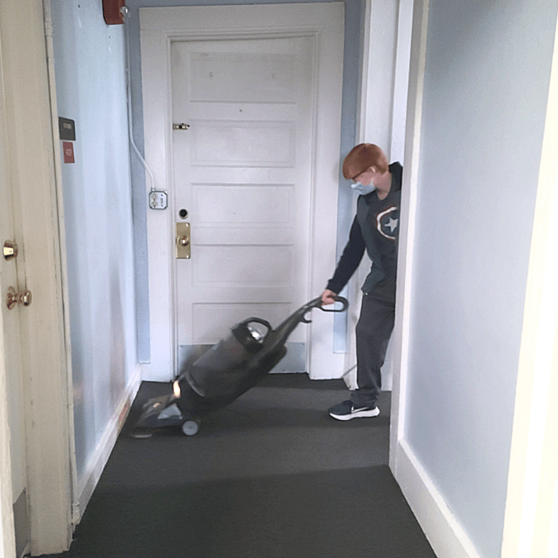Person in a mask vacuums a hallway. Redhead, wearing dark clothing. Black mat on floor, white door.