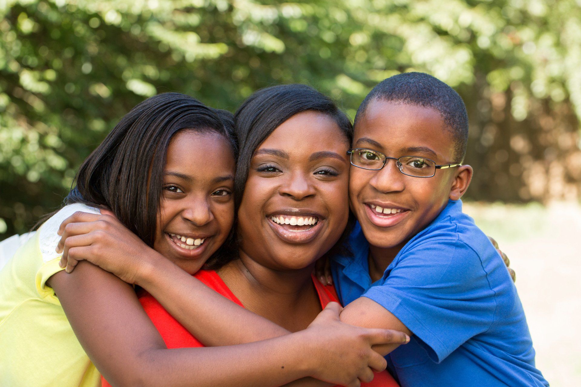 Woman hugs two smiling children outdoors.