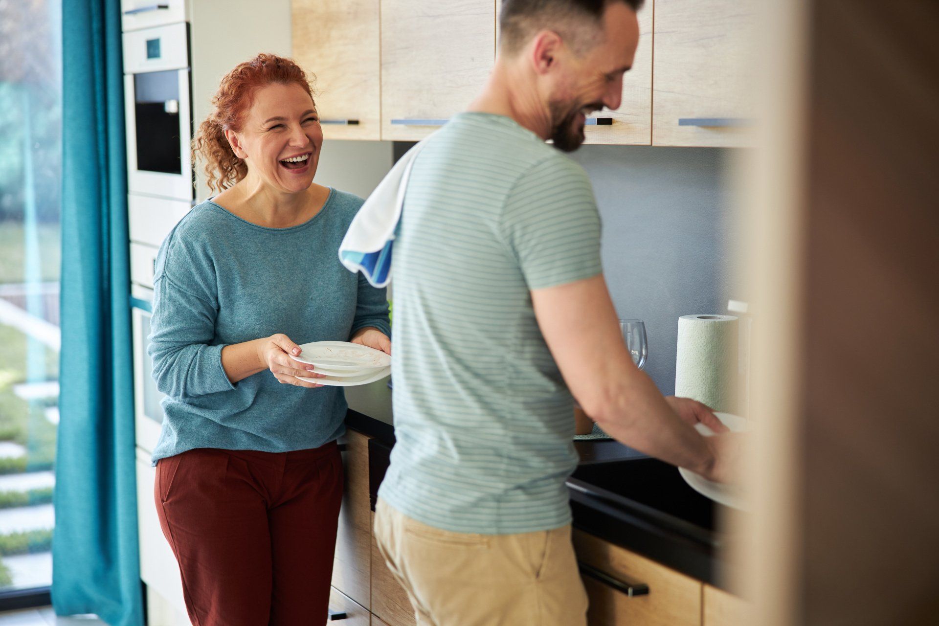 Woman laughs, holding plates; man washes dishes in kitchen.