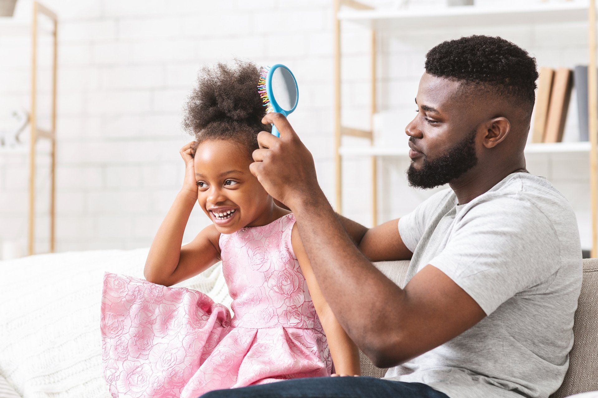 Father combing his daughter's hair, both smiling. They are indoors, daughter in pink dress, father in gray shirt.