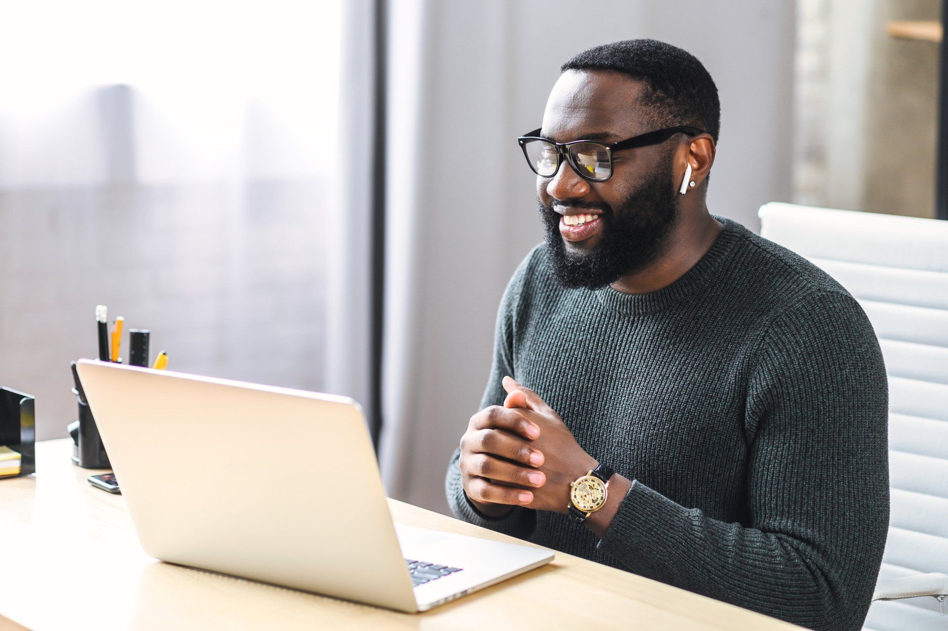 Man with glasses and a beard smiles while on a laptop video call. Indoors, at a desk.