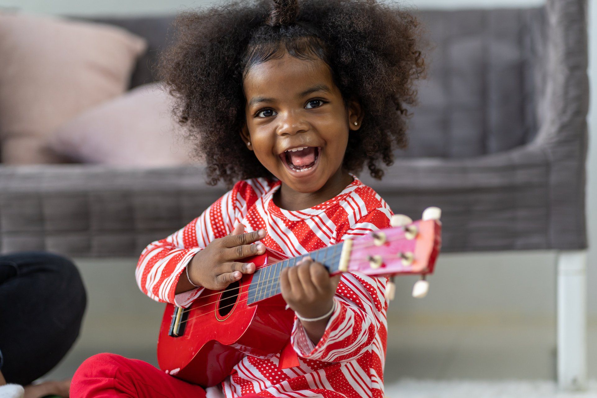 Young Black girl with curly hair, playing a red ukulele, smiling joyfully.