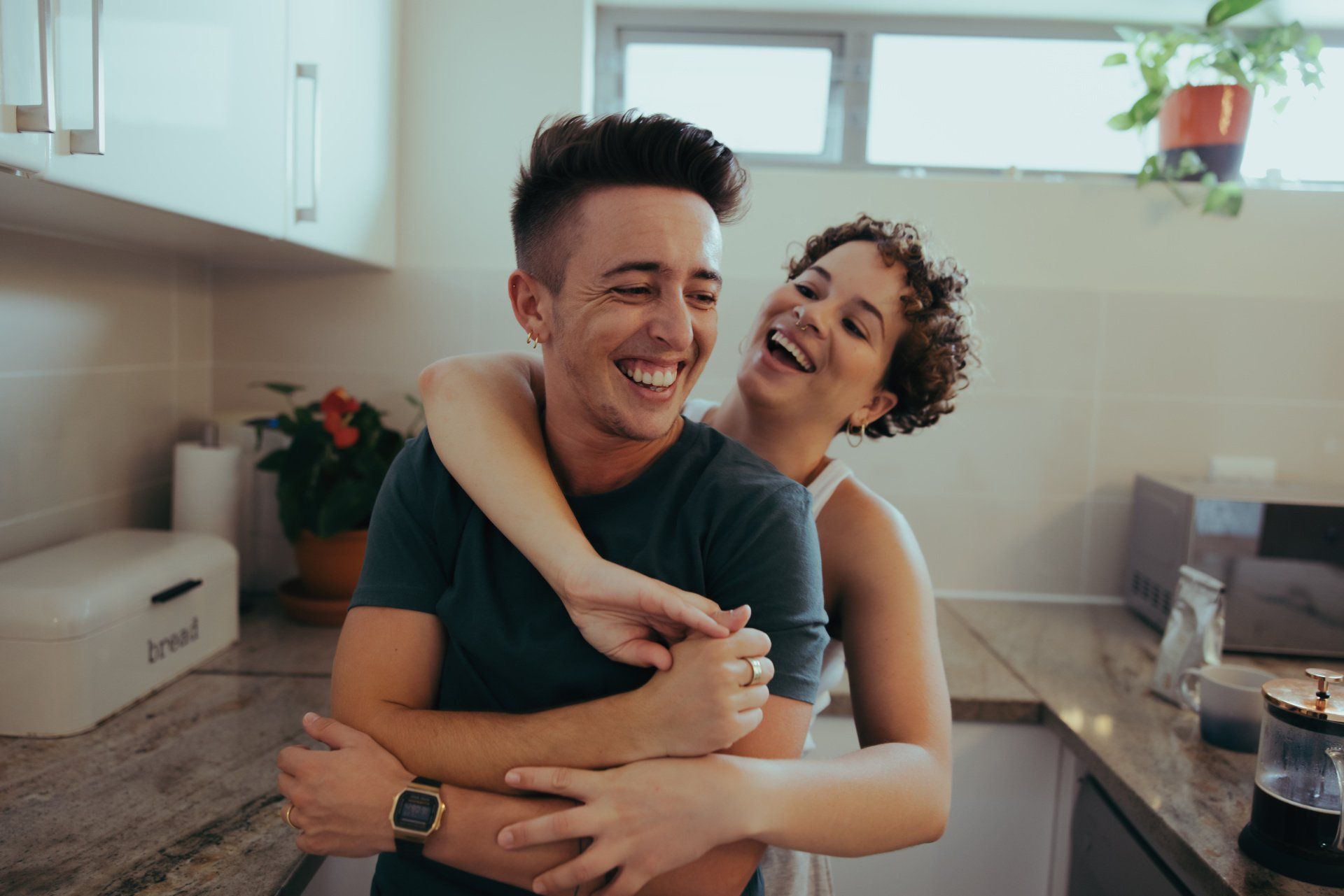 Woman hugging and laughing with a person in a kitchen; both smiling.