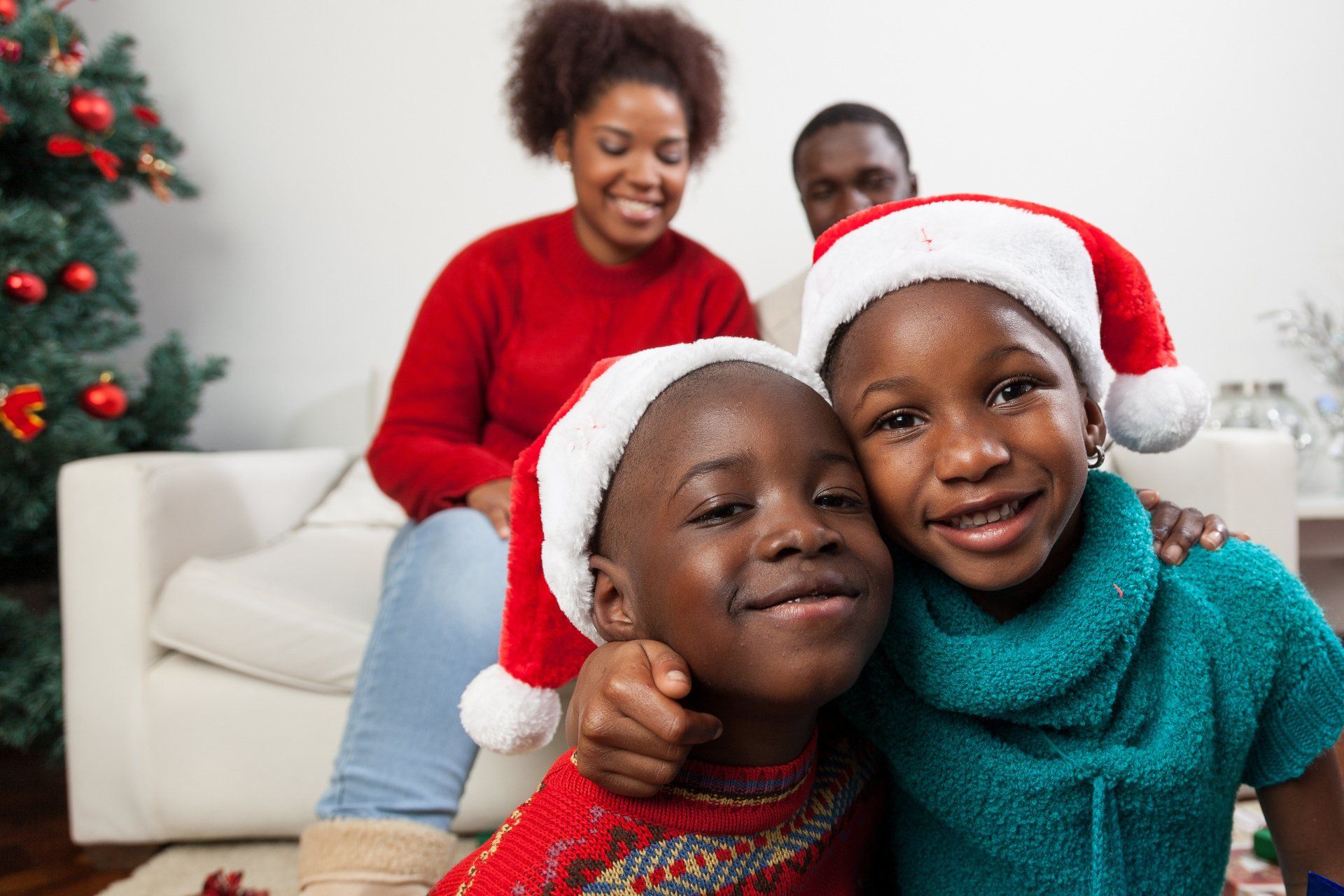 Two smiling children in Santa hats embrace; parents in background, Christmas tree visible.