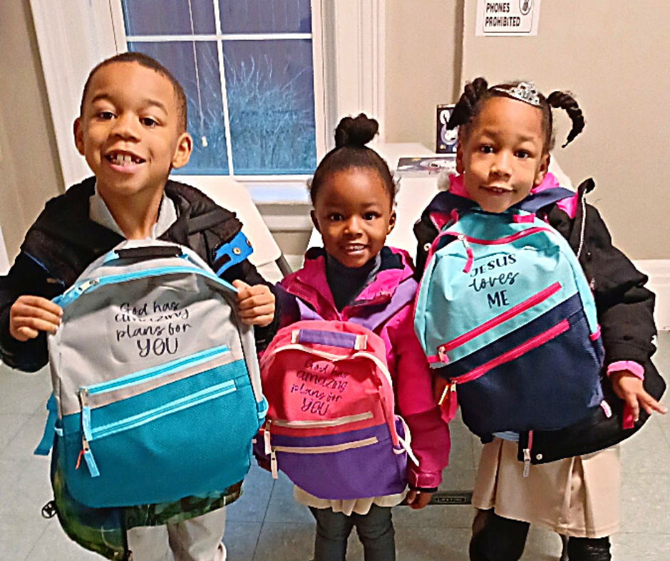 Three smiling children holding backpacks; two are turquoise and one is pink.