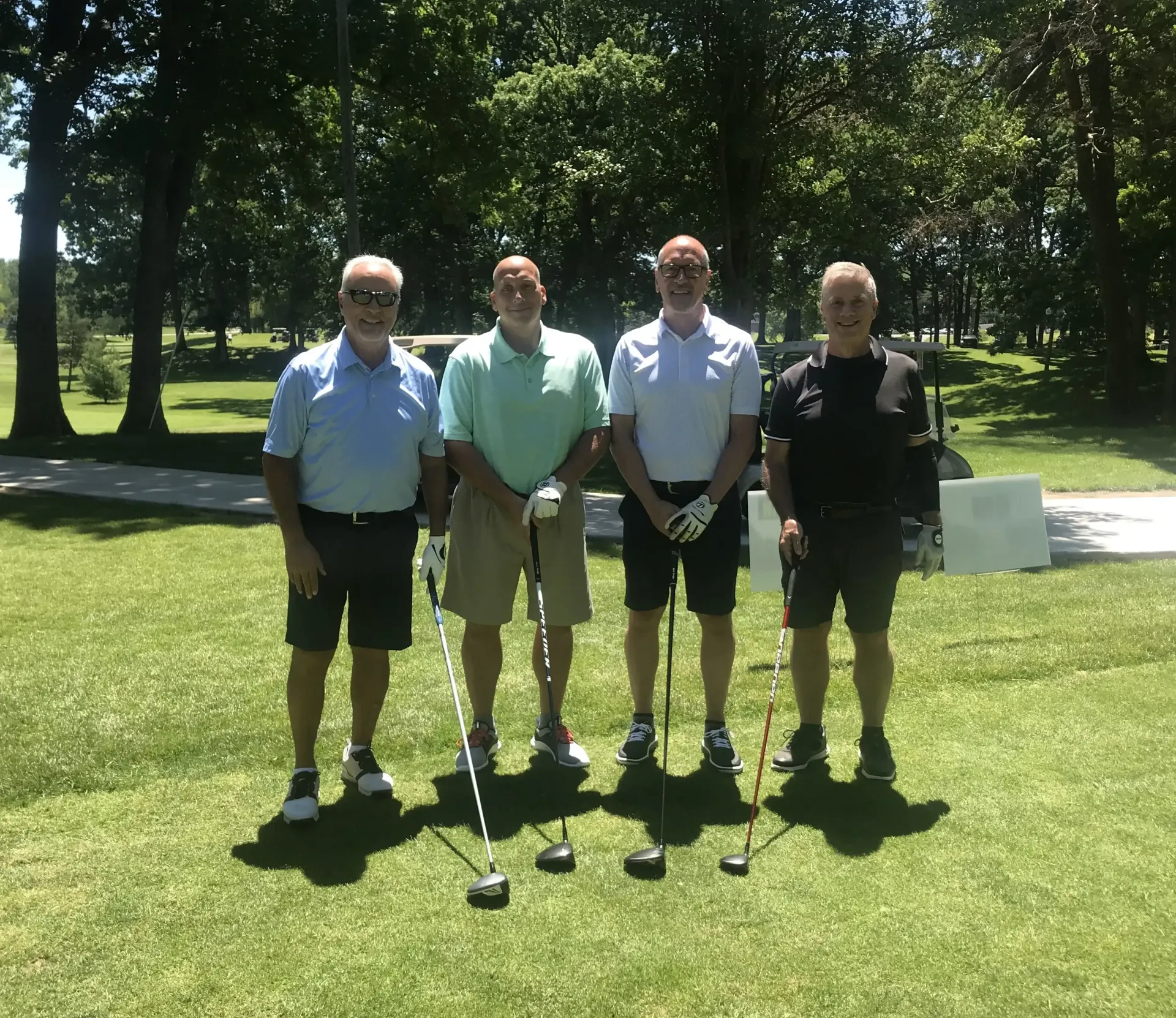 Four men standing on a golf course, holding golf clubs. They are wearing shorts and shirts on a sunny day.