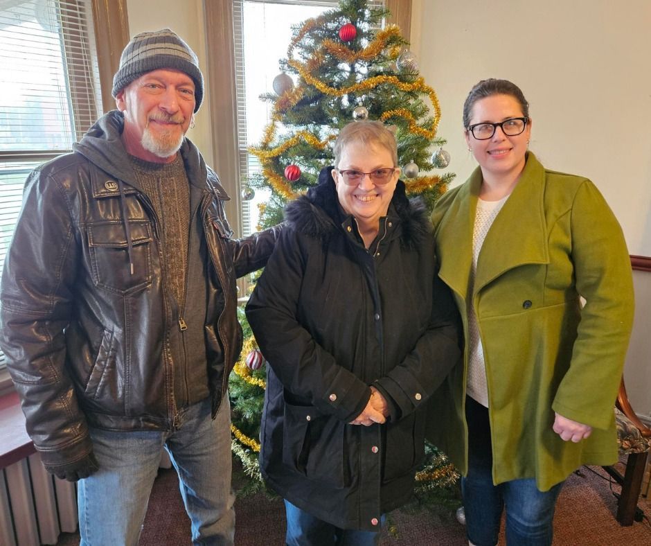 Three people stand by a decorated Christmas tree. Man wears a hat and jacket. Woman in coat smiles. Another woman in green coat.