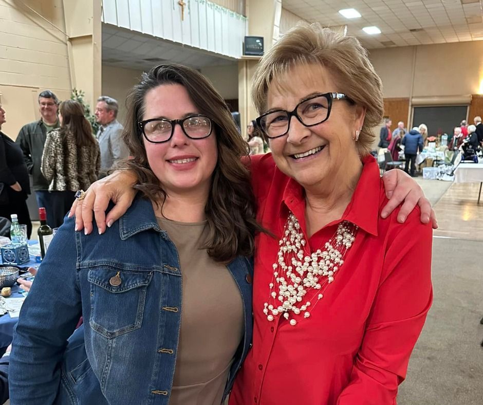 Two women smiling, one in denim jacket, one in red, arms around each other, indoors.