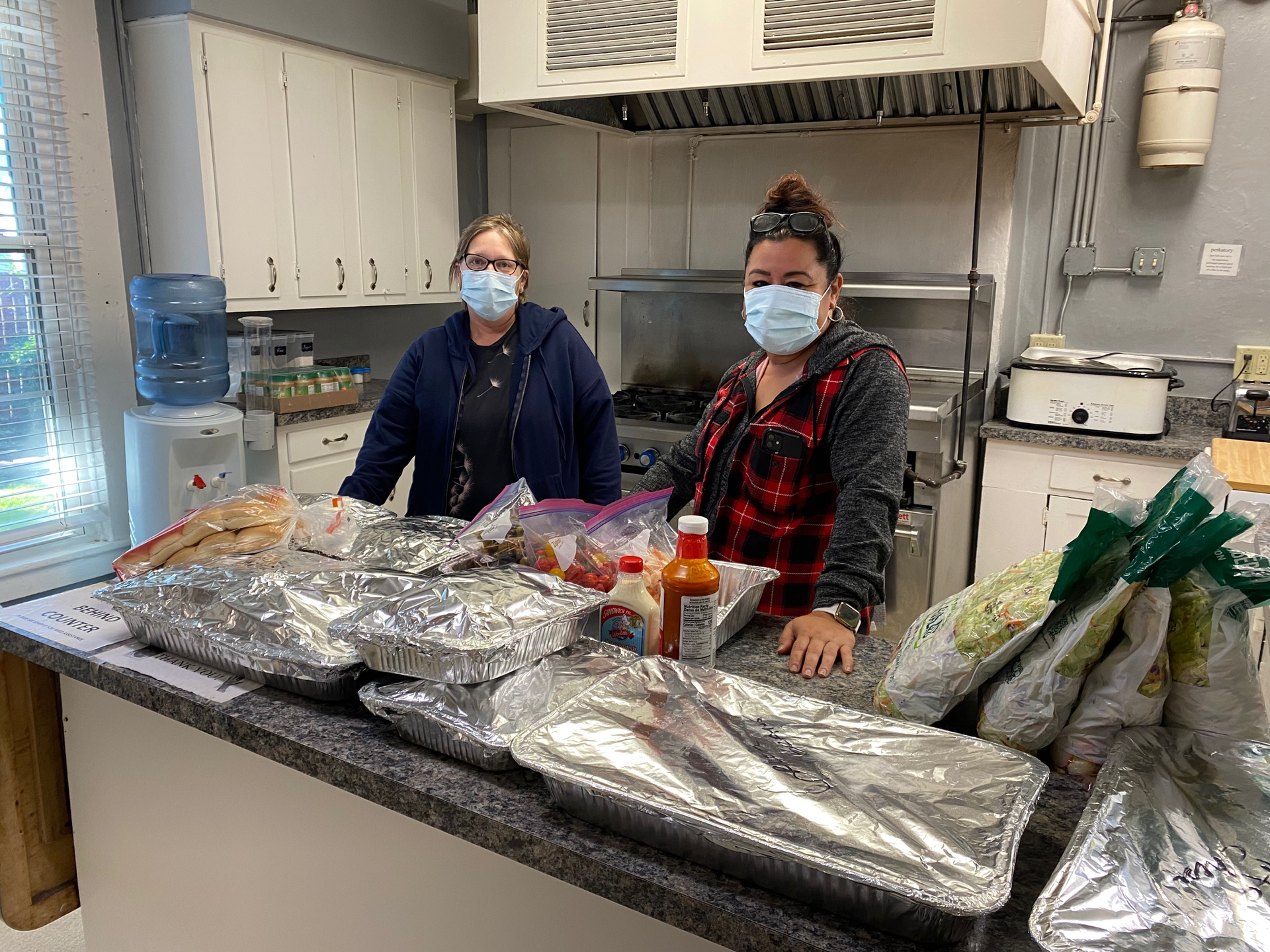 Two women wearing masks stand behind a counter with covered food containers in a kitchen.