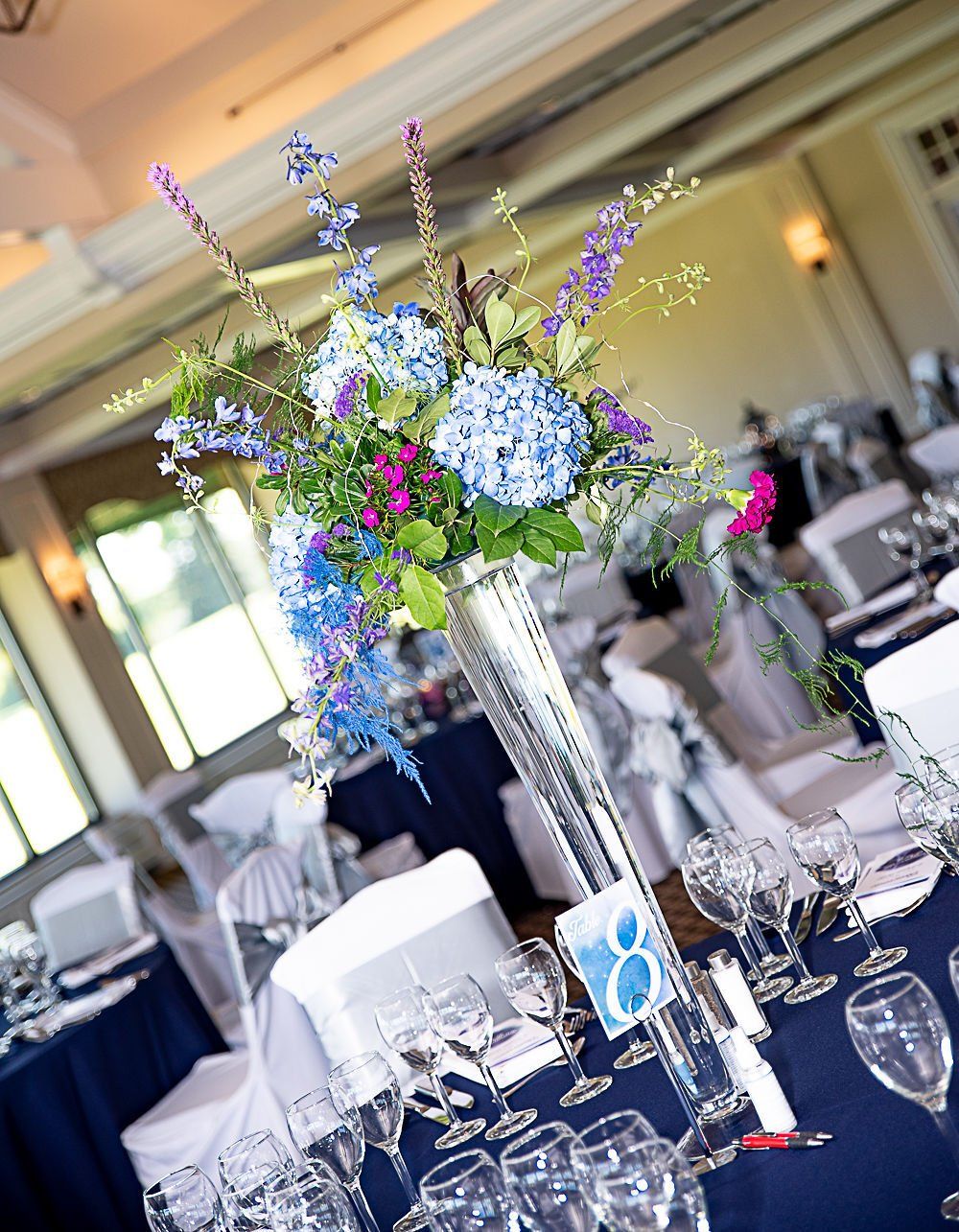 Floral centerpiece on a table set for a wedding, with blue and purple flowers, navy tablecloths, and white chairs.