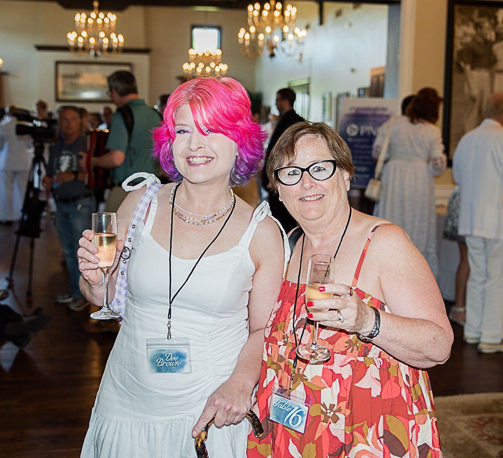 Two women smiling, holding drinks at an event. One has pink hair, the other wears glasses.