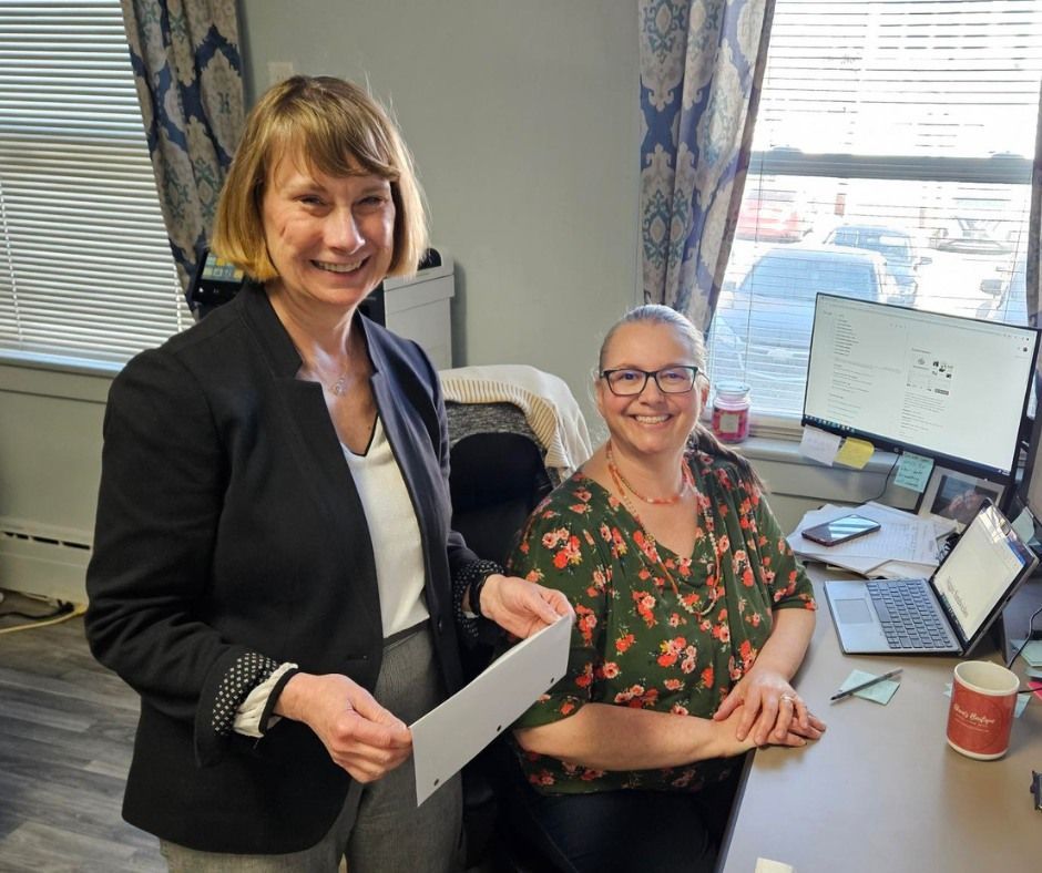 Two women smiling in an office; one holds papers, the other sits at a desk with a computer.