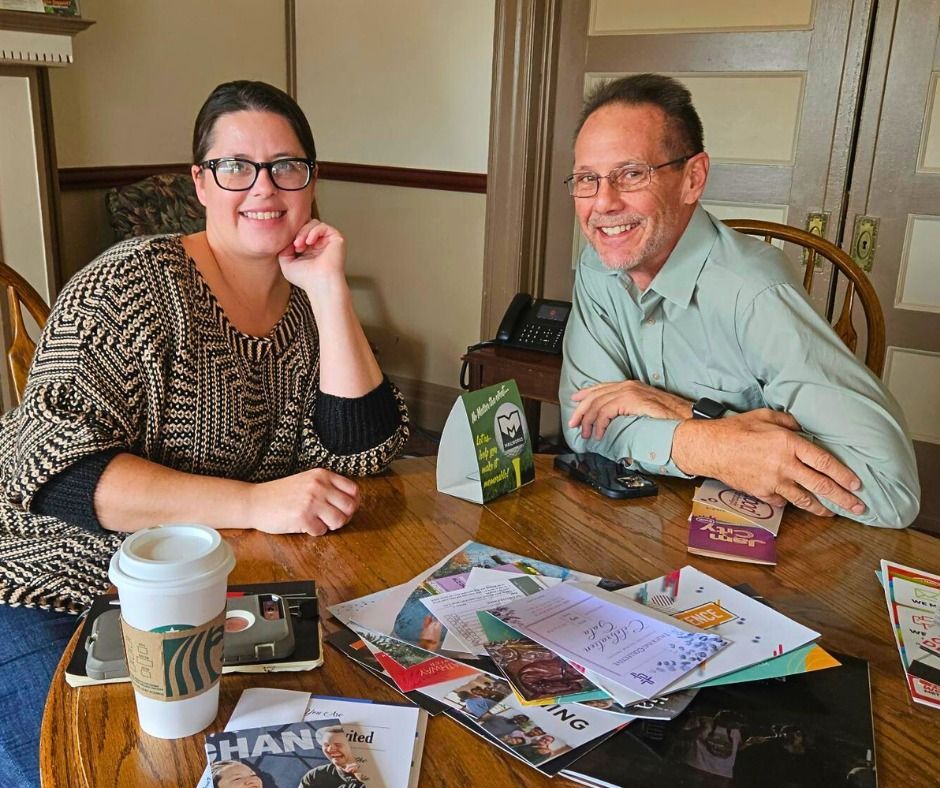Woman and man sit at table smiling. Papers, coffee cup, and phone visible. Wooden table in interior setting.