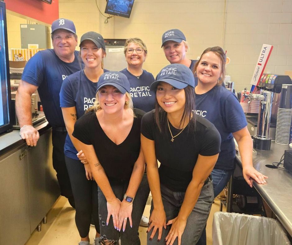 Group of seven Pacific Retail employees in blue shirts and hats smiling behind a counter.