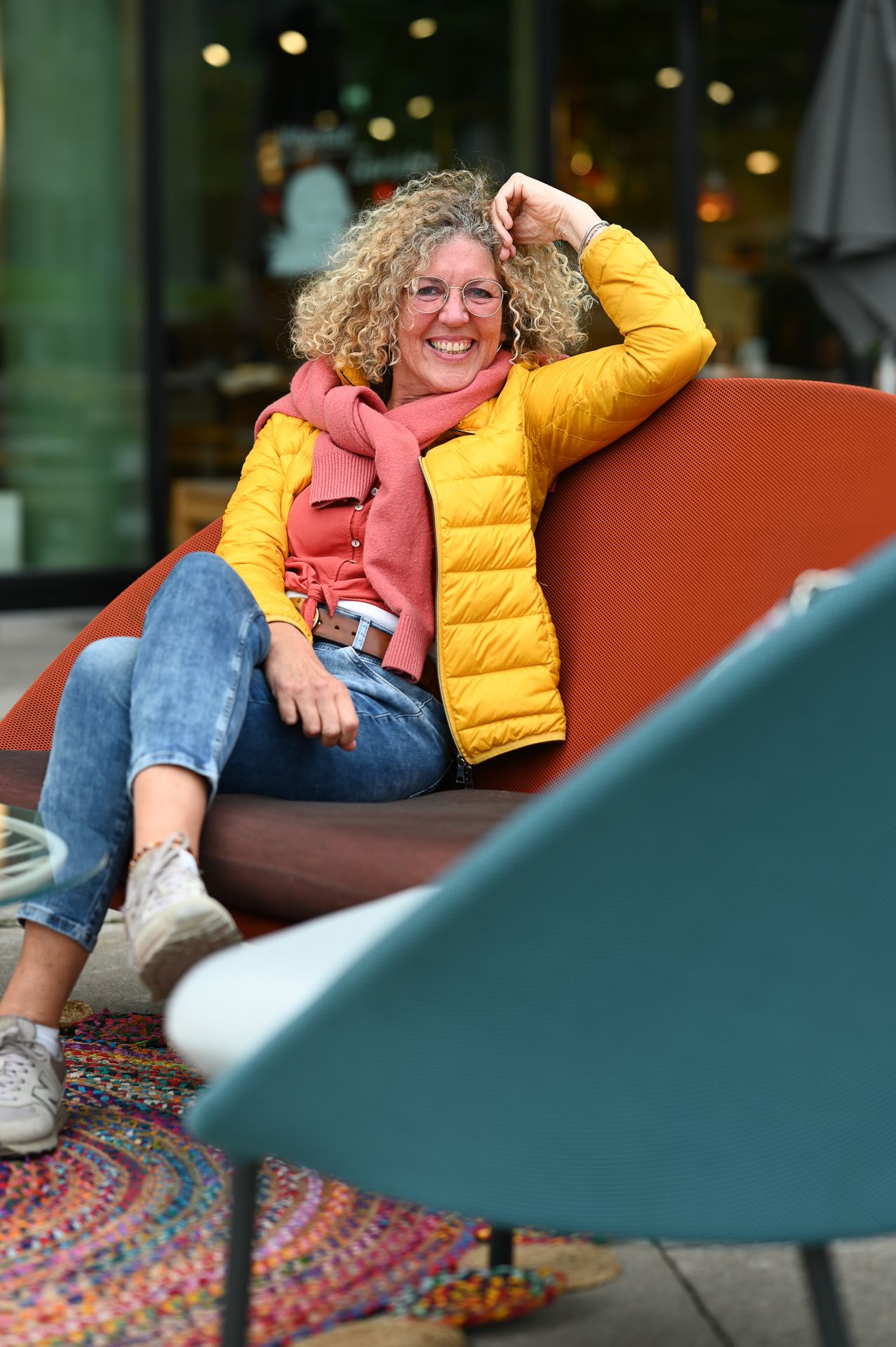 Eine Frau in einer gelben Jacke und einem rosa Schal sitzt lächelnd auf einem Liegestuhl auf der gemütlichen Caféterrasse