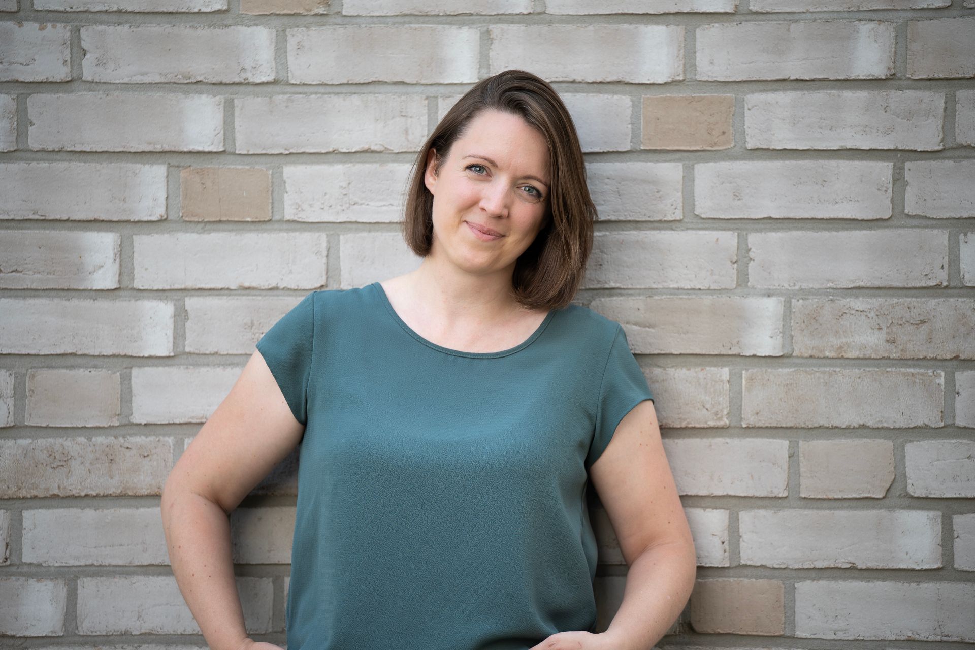 Eine Frau in einem türkisfarbenen Hemd lehnt lächelnd an einer Backsteinmauer.