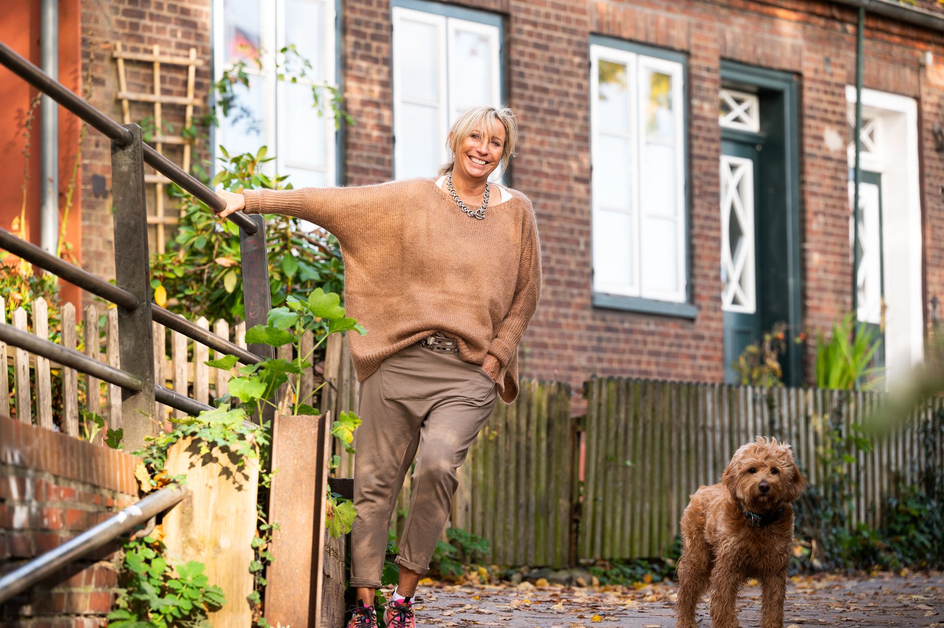 Frau lächelt, lehnt an einem Geländer neben einem braunen Hund, im Hintergrund ein Backsteingebäude, trägt einen braunen Pullover und eine braune Hose.