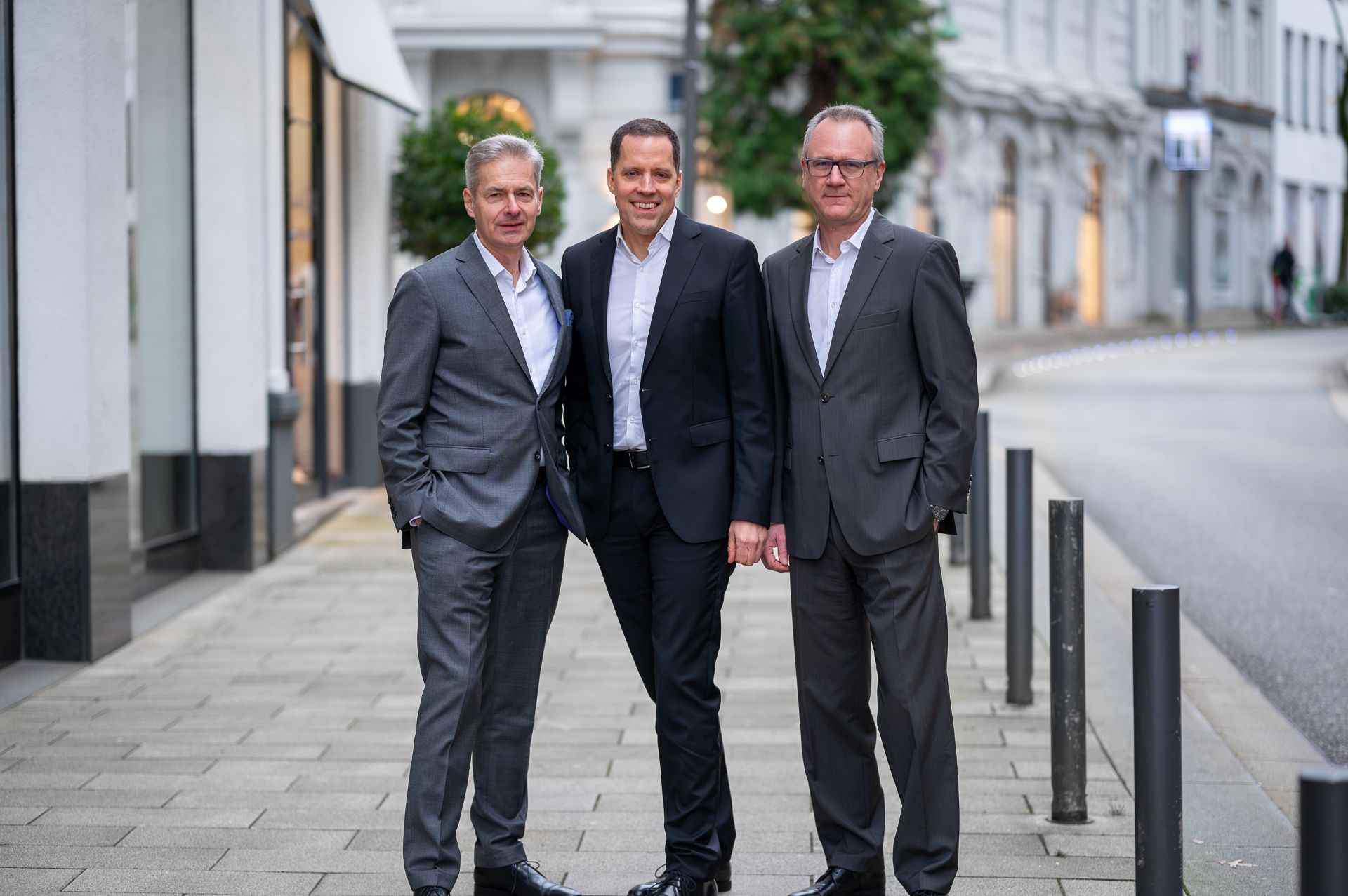 Drei Männer in Anzügen stehen lächelnd auf einem Bürgersteig; im Hintergrund sind Straße und Gebäude zu sehen.