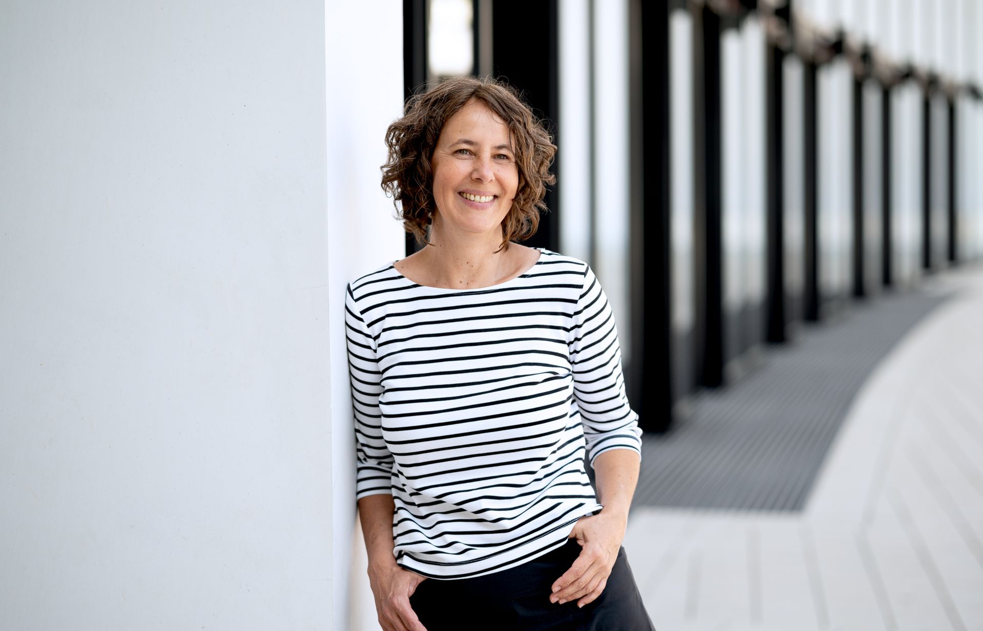 Frau mit lockigem Haar, in gestreiftem Hemd, lehnt lächelnd an einer weißen Wand. Im Hintergrund ein modernes Gebäude.