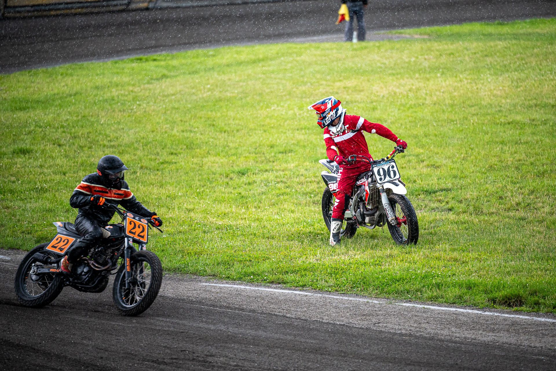 Motorrad-Flat-Track-Rennfahrer auf nasser Strecke. Ein Fahrer in Orange, ein anderer in Rot, in der Nähe einer Grasfläche.