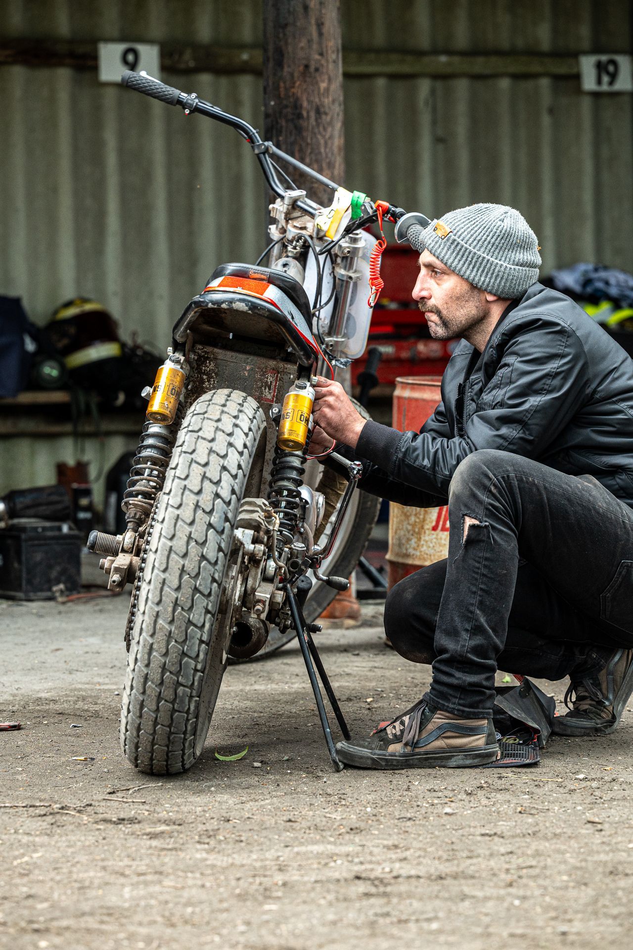 Ein Mann hockt neben einem Motorrad und justiert es. Er trägt eine Mütze, eine schwarze Jacke und befindet sich in einer Garage.