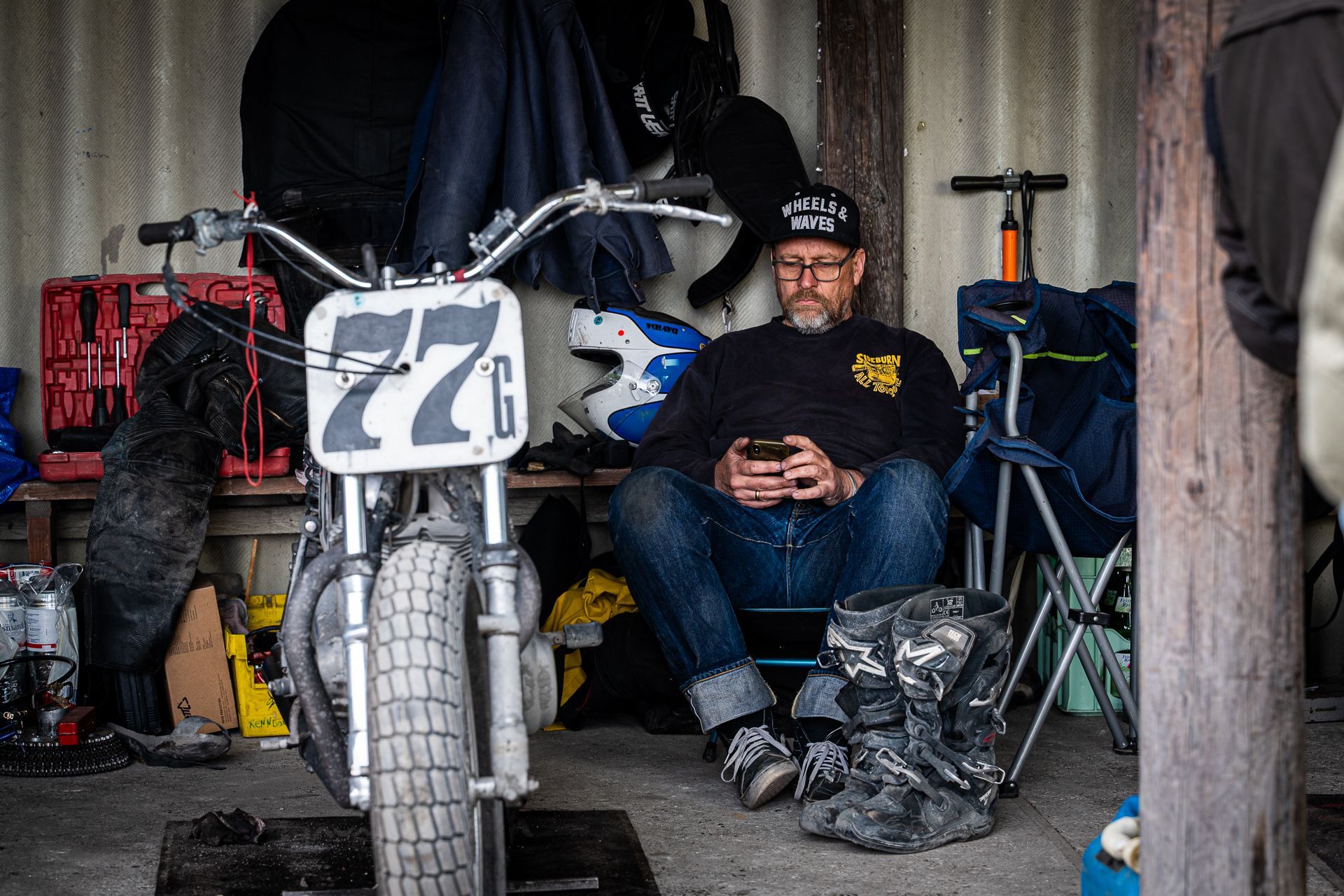 Ein Mann telefoniert und sitzt neben einem Flat-Track-Motorrad (77) in einer provisorischen Garage; Werkzeug und Ausrüstung liegen verstreut herum.