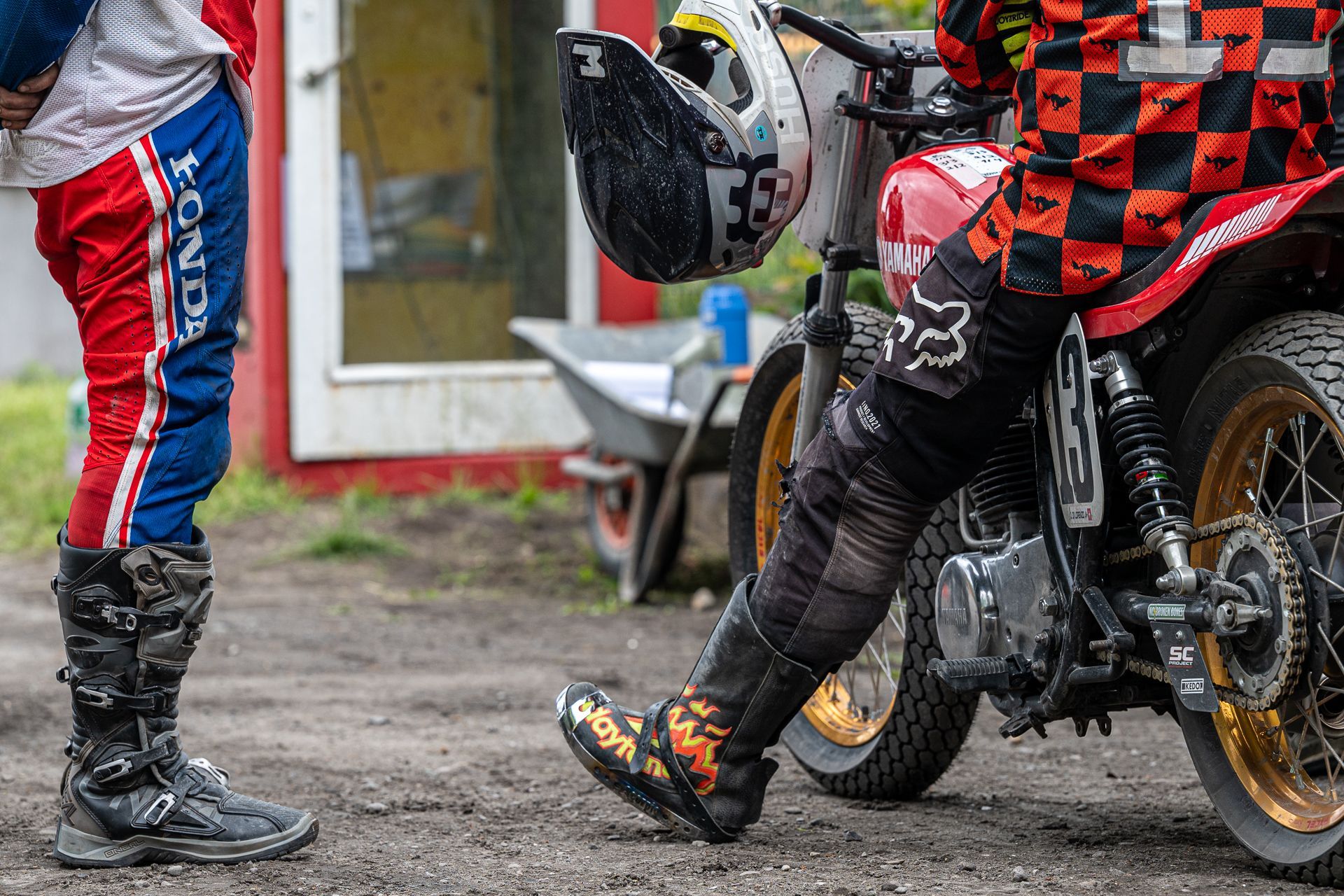 Zwei Personen in Motocross-Ausrüstung; eine auf einem Motorrad, die andere daneben stehend. Farben: Rot, Blau und Schwarz. Freiluftaufnahme.