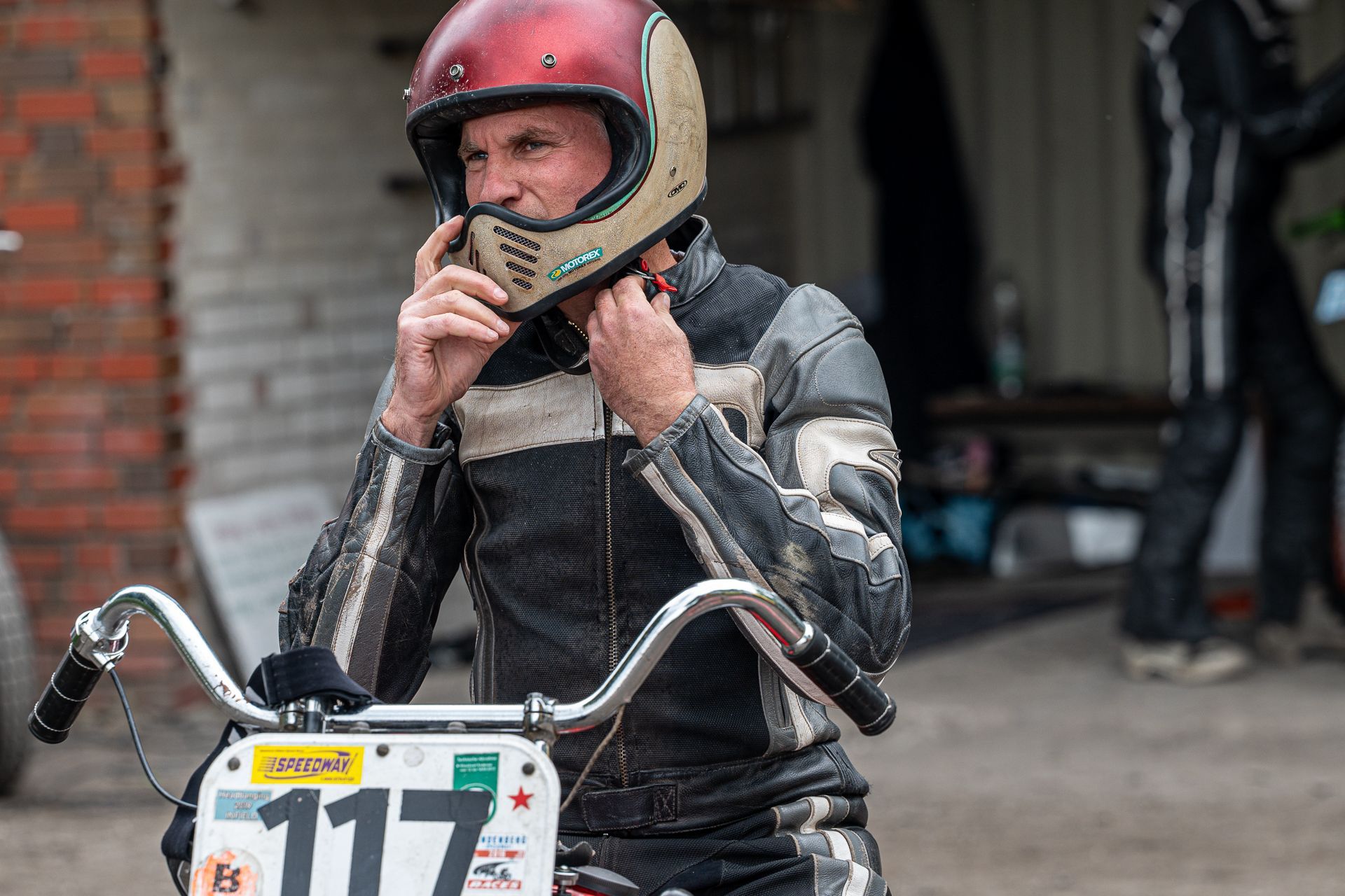 Mann in Lederjacke und Helm justiert den Helmriemen an einem Oldtimer-Motorrad, im Freien.