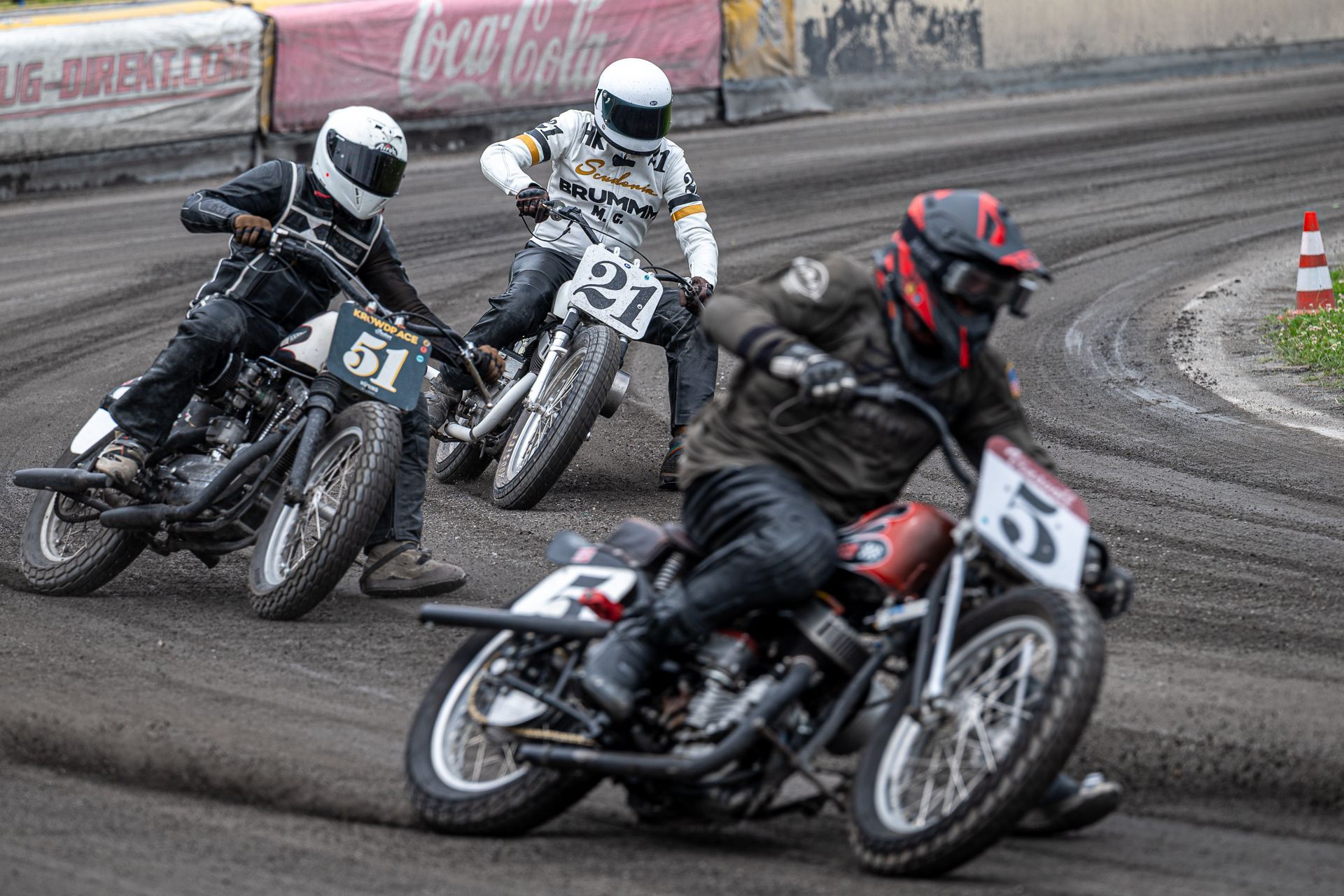Drei Motorradrennfahrer auf einer unbefestigten Rennstrecke, die mit hoher Geschwindigkeit durch eine Kurve fahren.
