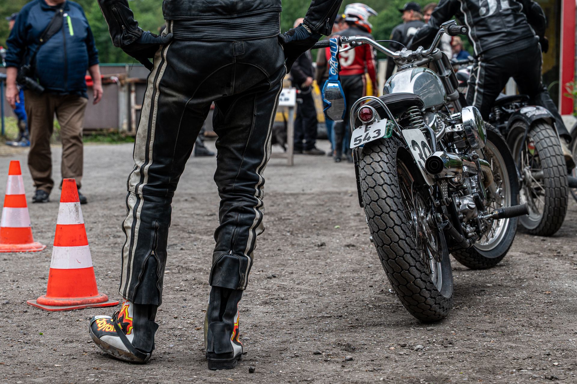 Ein Motorradfahrer in schwarzer Lederkluft steht mit erhobenen Armen neben einem Motorrad; im Hintergrund sind weitere Personen und Pylonen zu sehen.