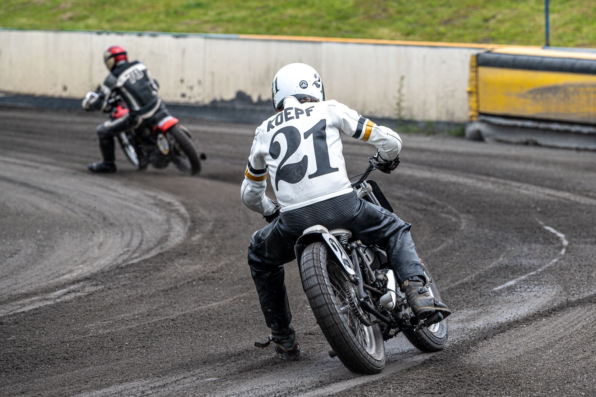 Zwei Motorradrennfahrer auf einer unbefestigten Rennstrecke, einer in Weiß mit der Nummer 21. Sie legen sich in eine Kurve.