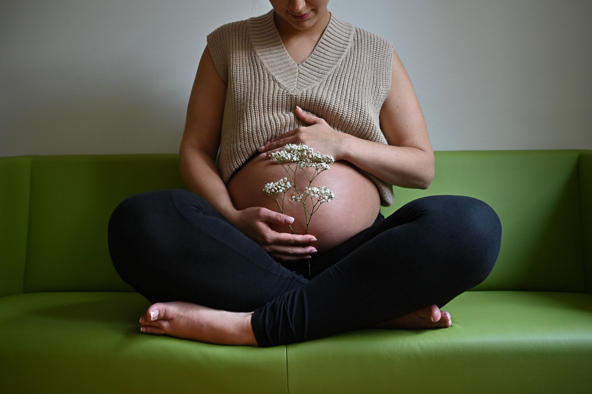 Schwangere sitzt im Schneidersitz auf einem grünen Sofa, die Hände mit Blumen auf dem Bauch.