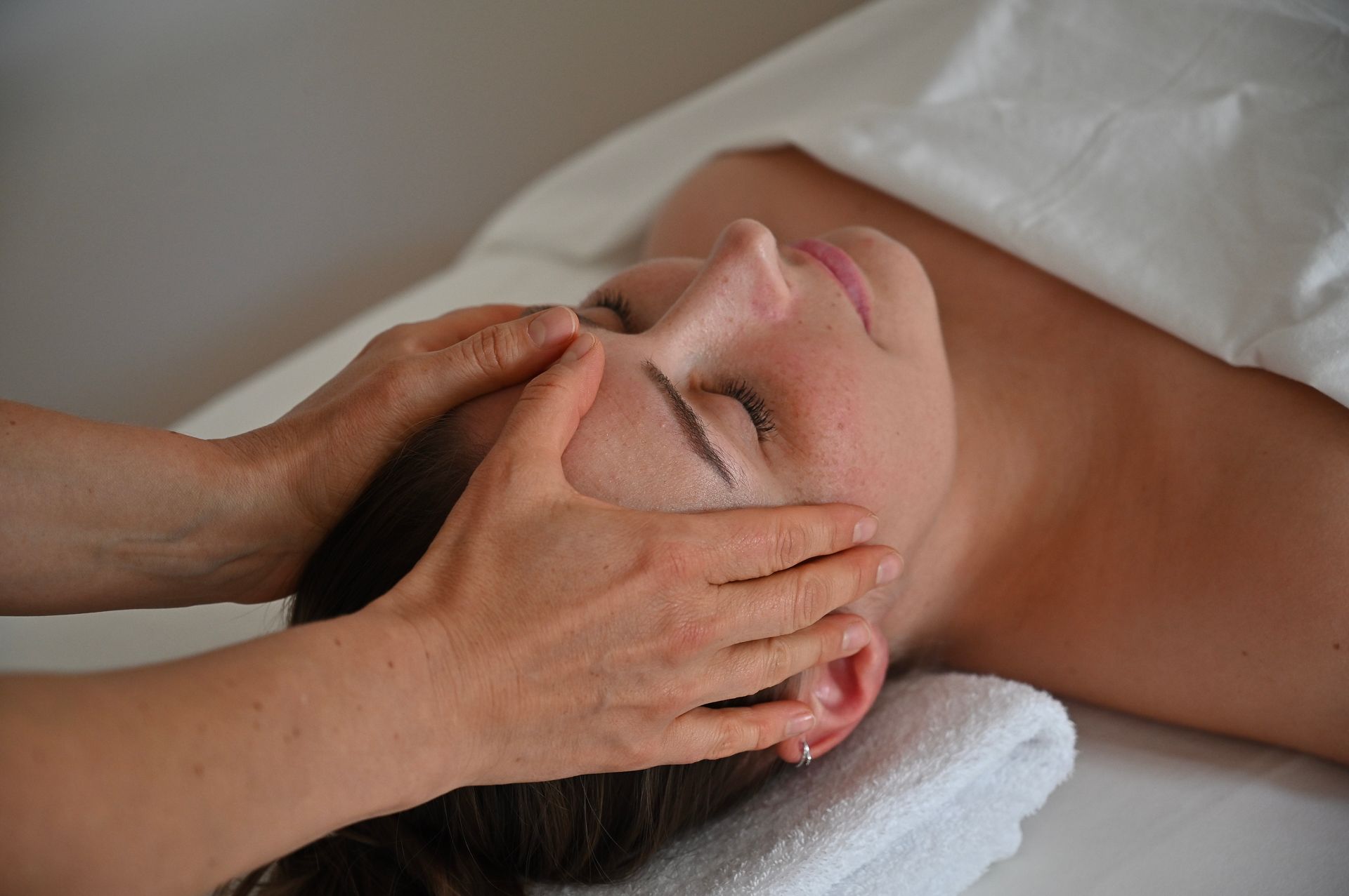 Person erhält eine Gesichtsmassage; Hände im Gesicht, liegt auf einem Handtuch, weiße Bettwäsche.