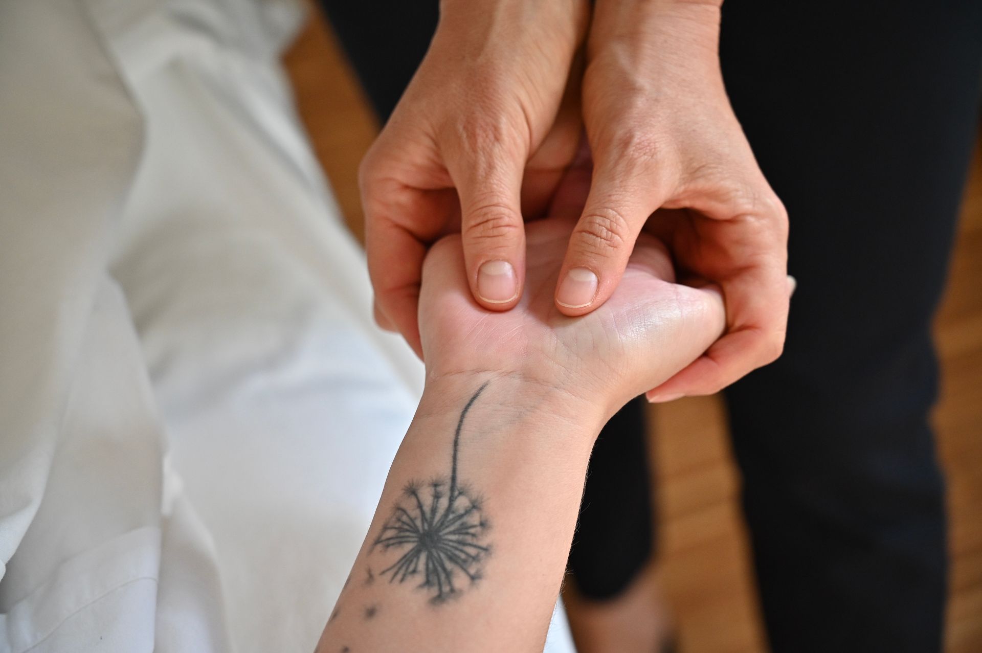 Hände massieren ein Handgelenk mit einem Löwenzahn-Tattoo, das auf einem weißen Stoff ruht.