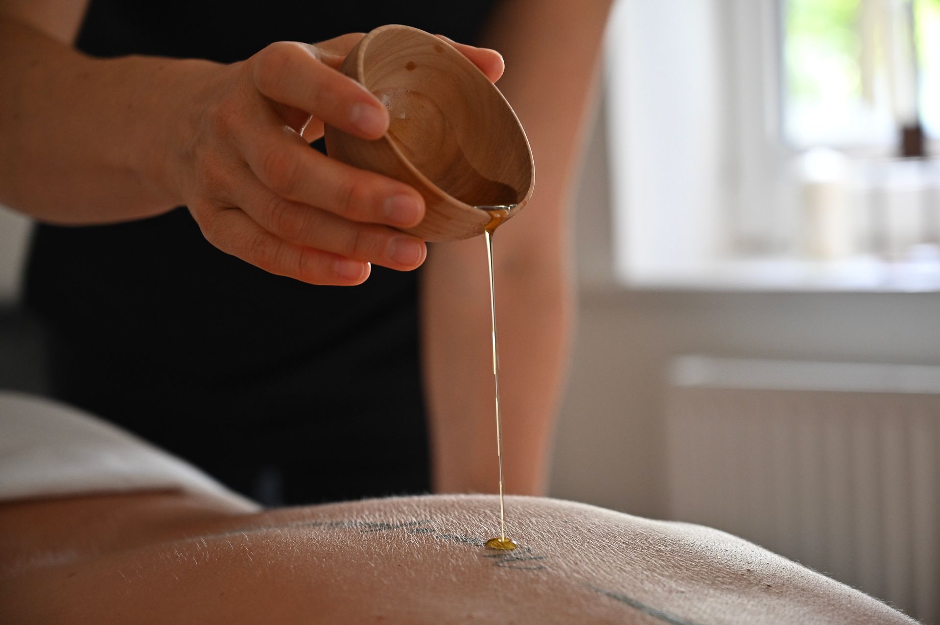 Eine Person gießt während einer Massage Öl aus einer Holzschale auf den Rücken einer anderen Person.