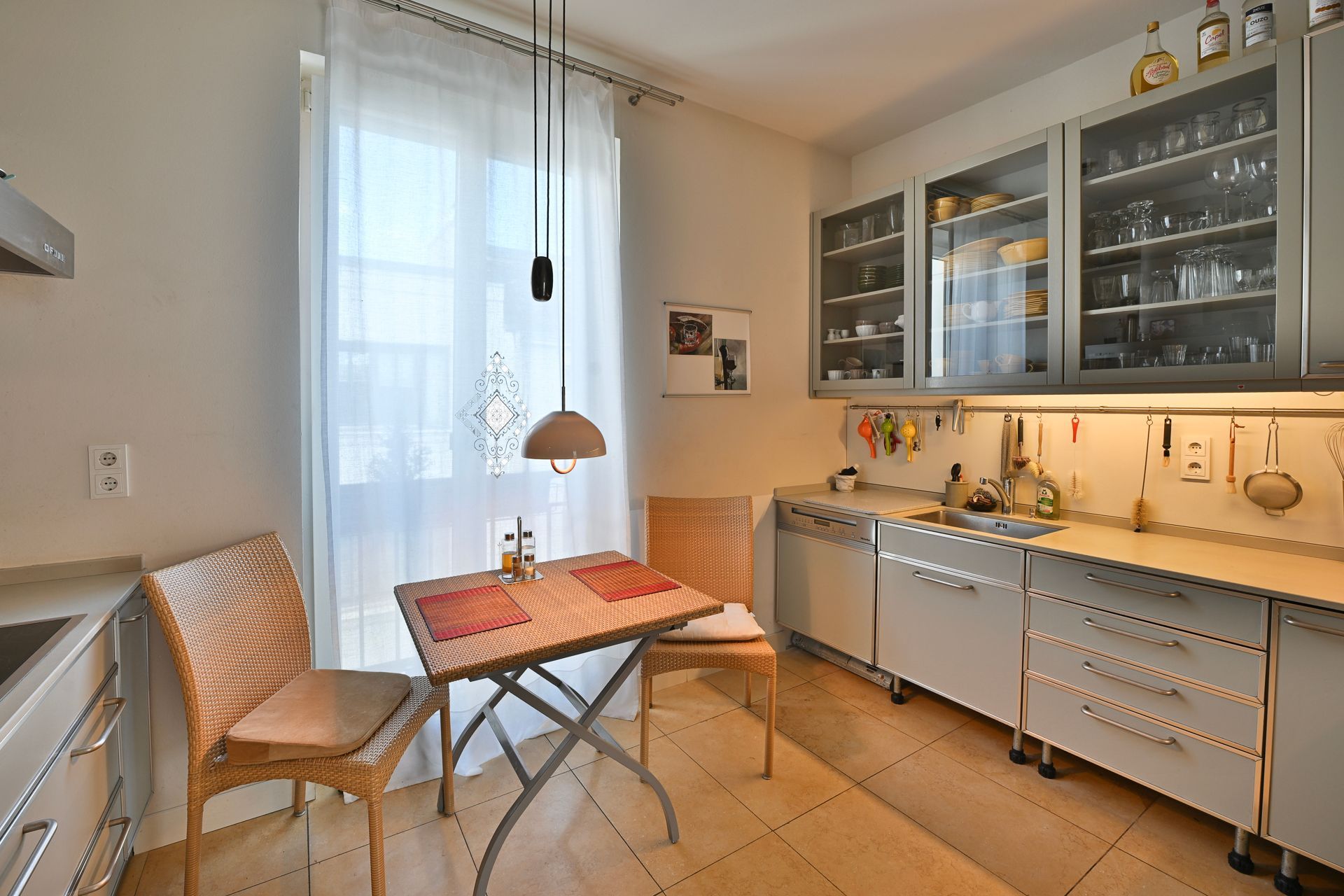 Küche mit Tisch und Stühlen am Fenster, weißen Schränken und Deckenbeleuchtung.