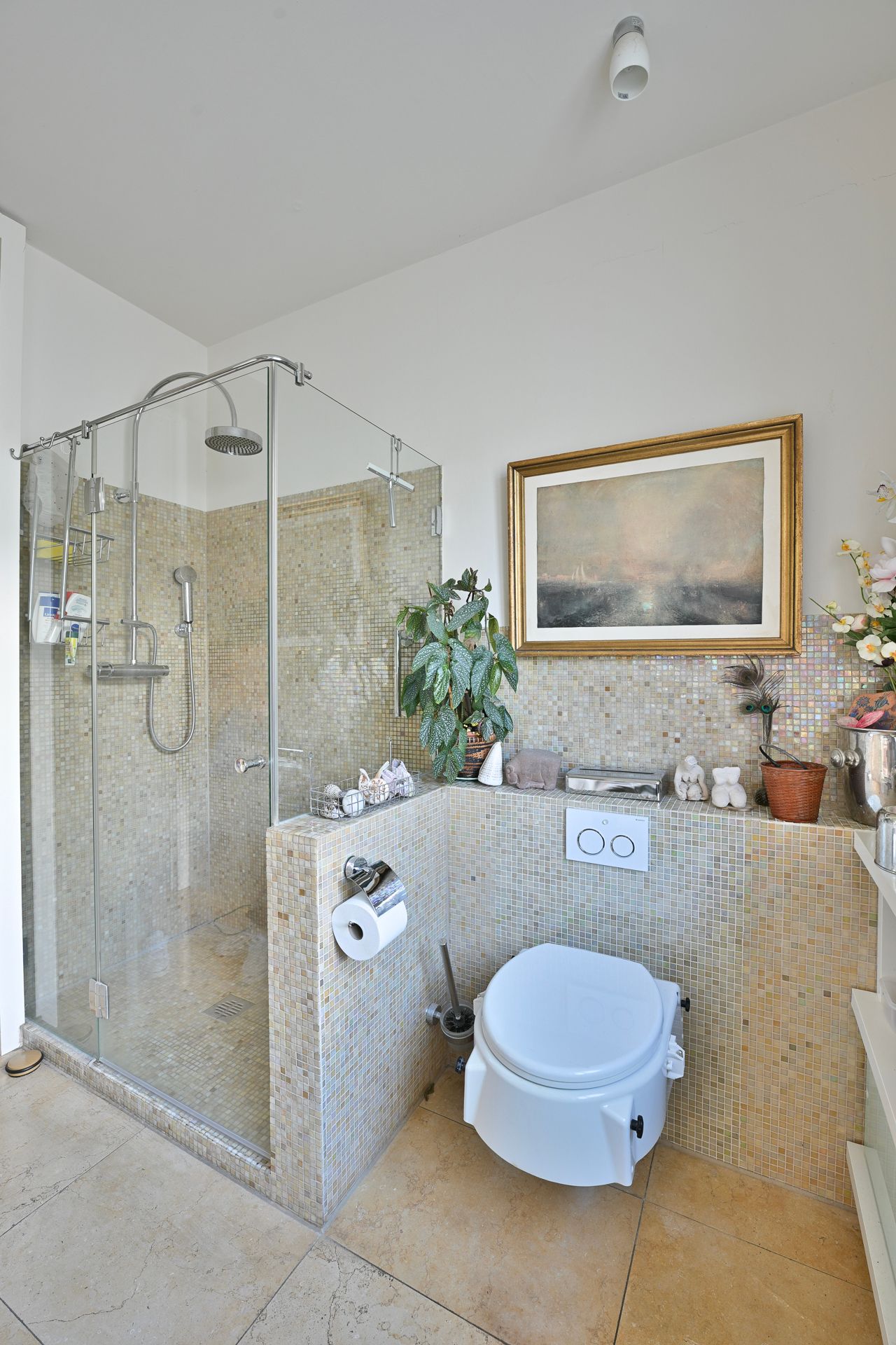 Badezimmer mit Mosaikfliesen an Wänden und Dusche, WC, Kunstwerken. Helle Farbgestaltung.
