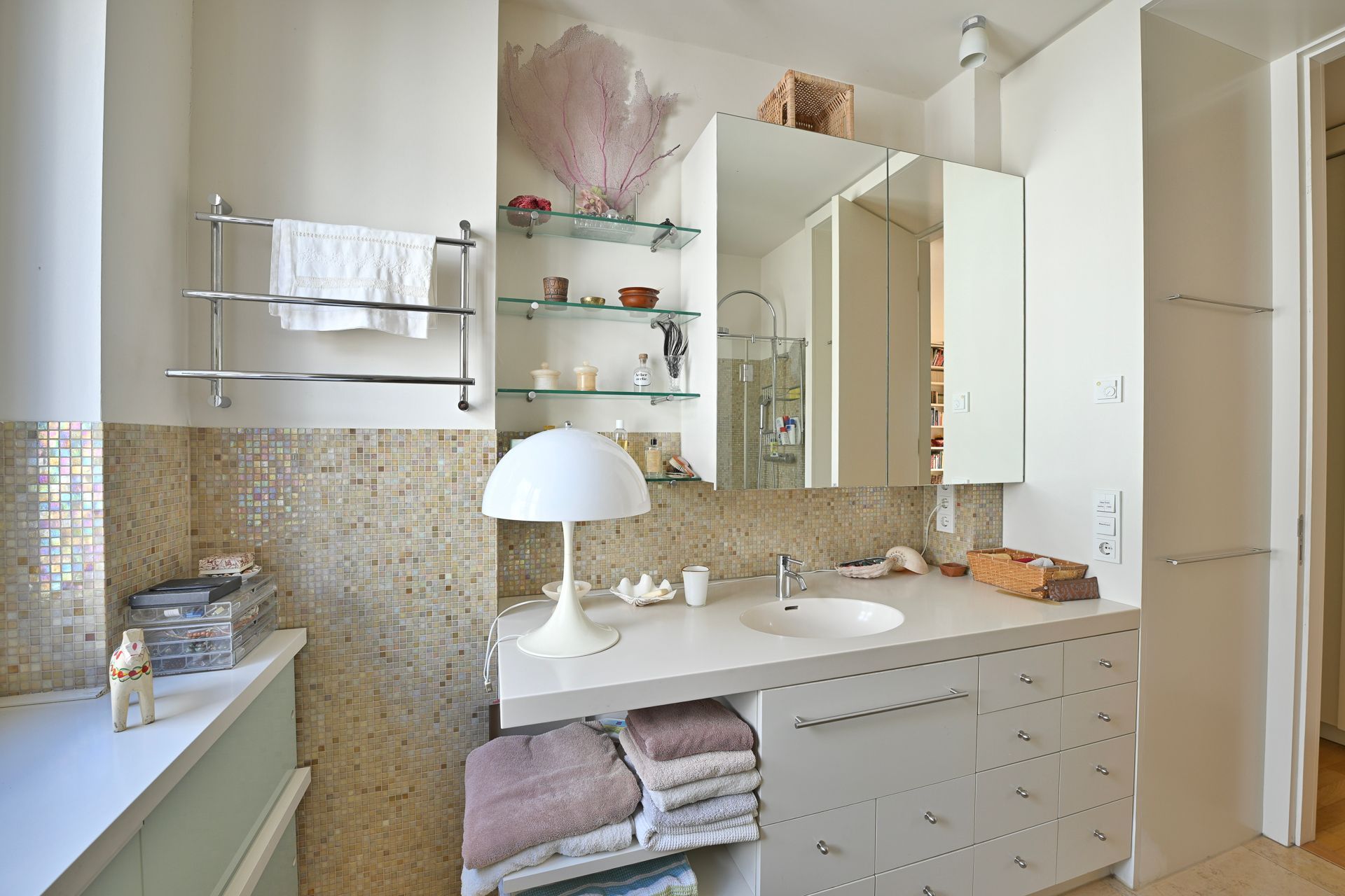 Badezimmer mit weißem Waschtisch, Mosaikfliesen und Glasregalen zur Aufbewahrung von Toilettenartikeln.