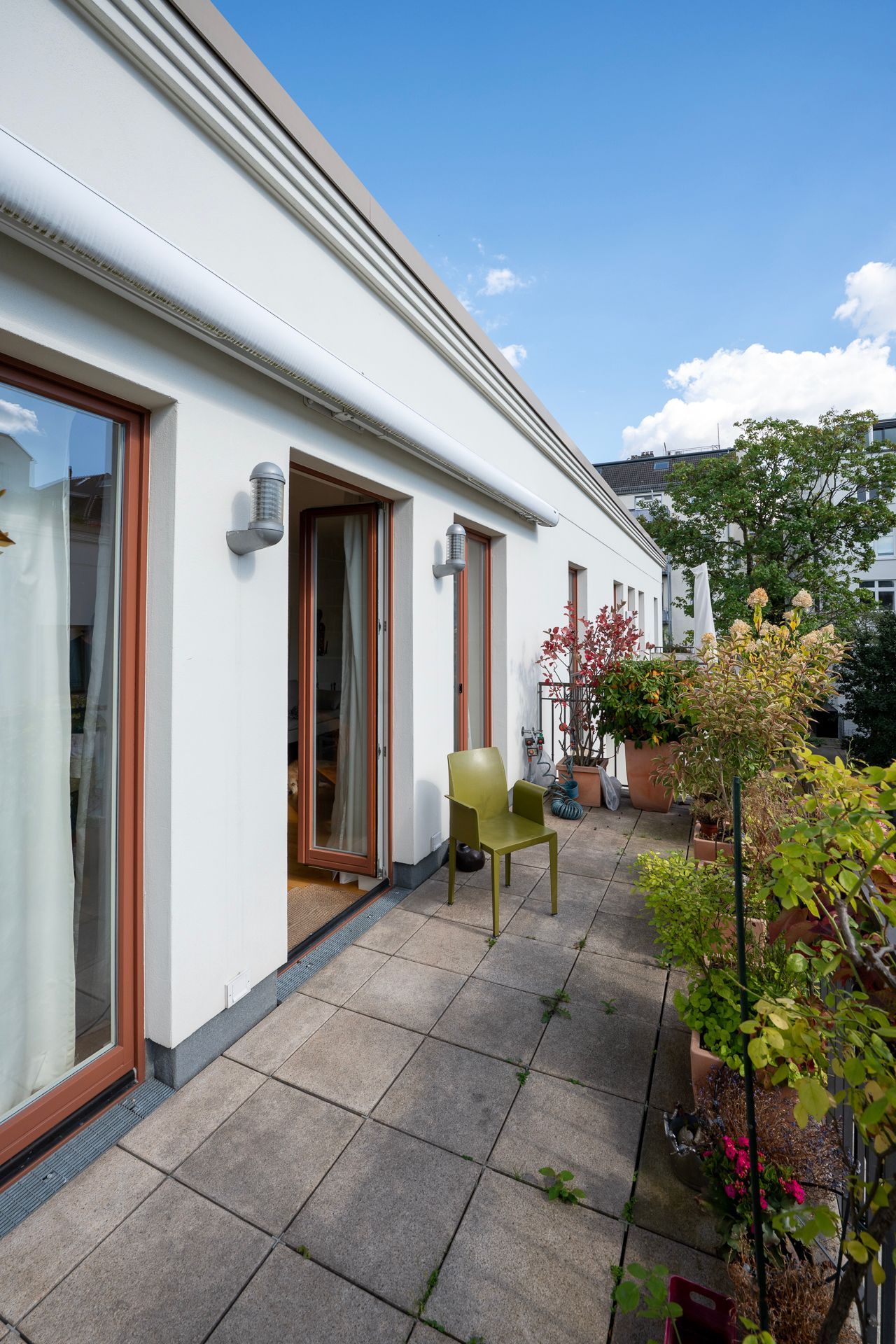 Balkon mit einem grünen Stuhl, Topfpflanzen und offenen Türen. Sonniger Tag.