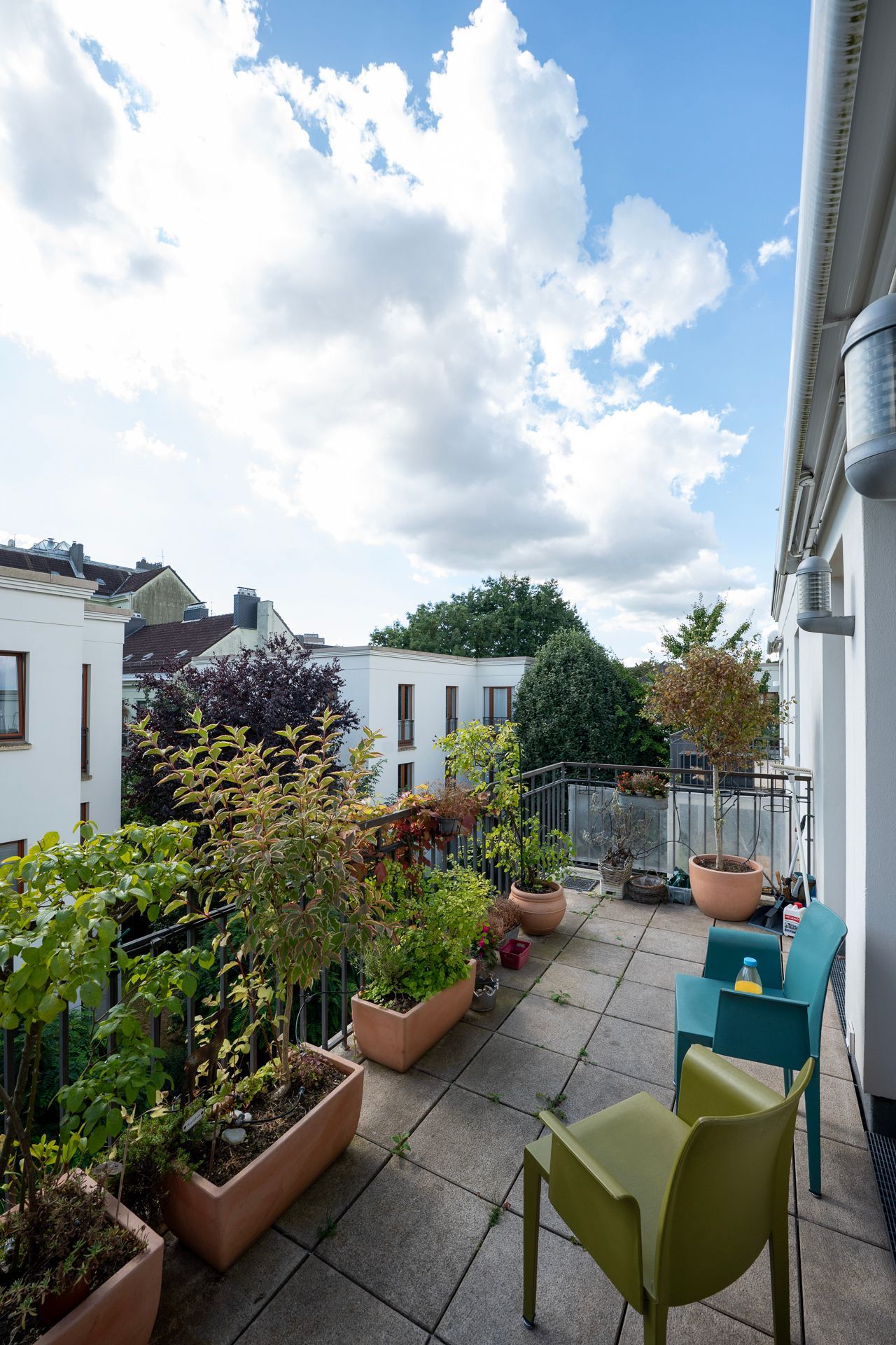 Balkon mit Topfpflanzen, bunten Stühlen und einem bewölkten blauen Himmel. Im Hintergrund sind Gebäude zu sehen.