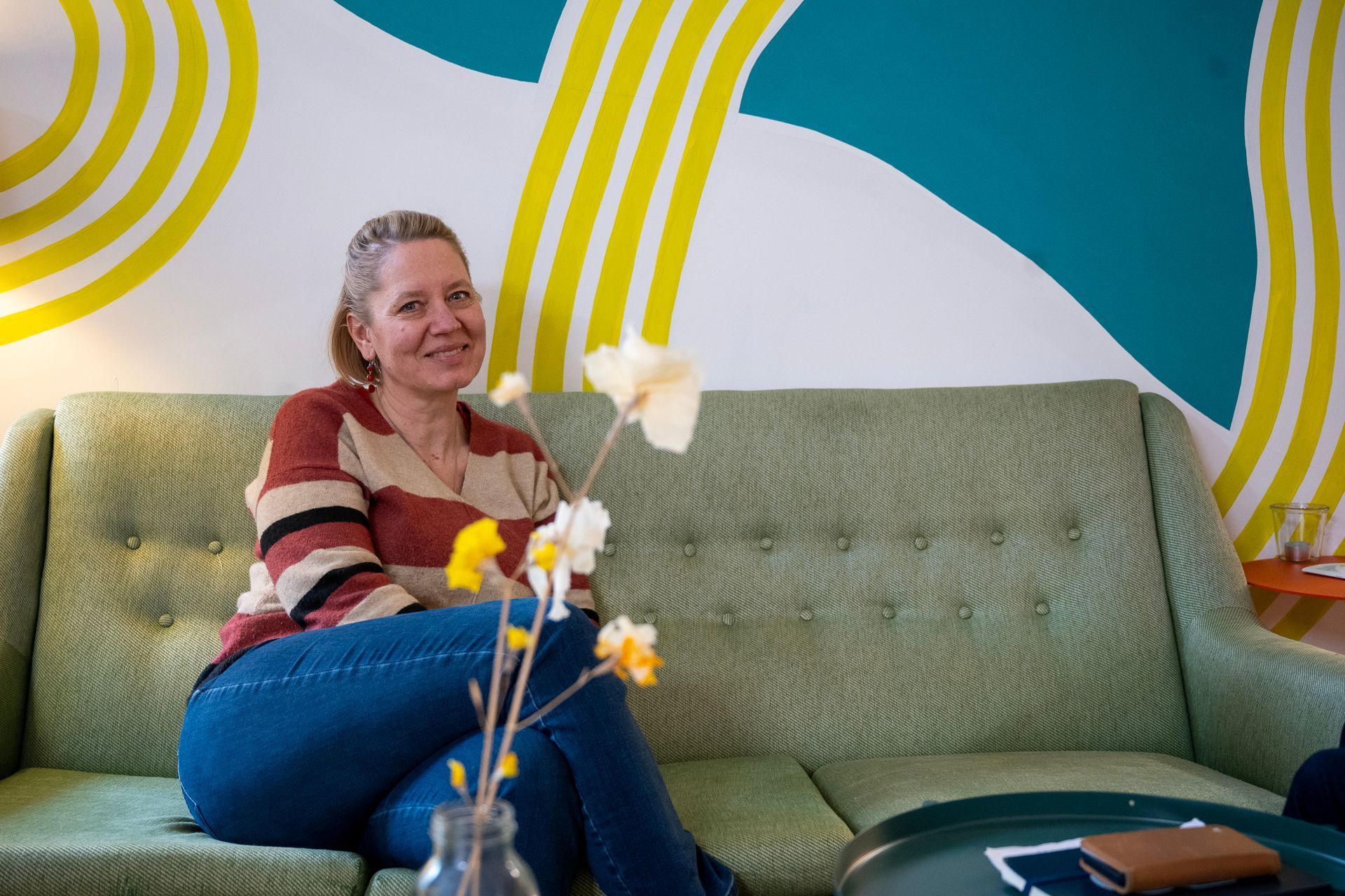 Frau sitzt lächelnd auf einem grünen Sofa, Blumen im Vordergrund, farbenfrohe Wandmalerei im Hintergrund.