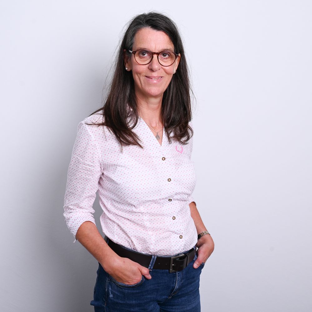 Frau mit Brille, rosa Hemd und Jeans lächelt, die Hände in den Taschen, vor einer weißen Wand.