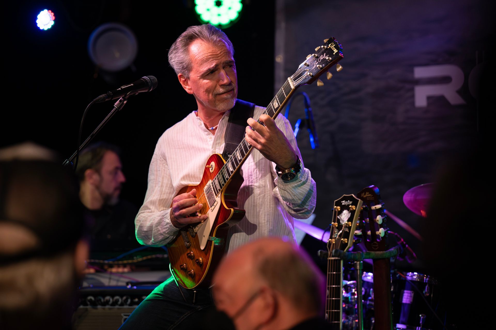 Ein Gitarrist spielt auf der Bühne; er trägt ein weißes Hemd. Bühnenbeleuchtung setzt ihn und die Gitarren im Hintergrund in Szene.