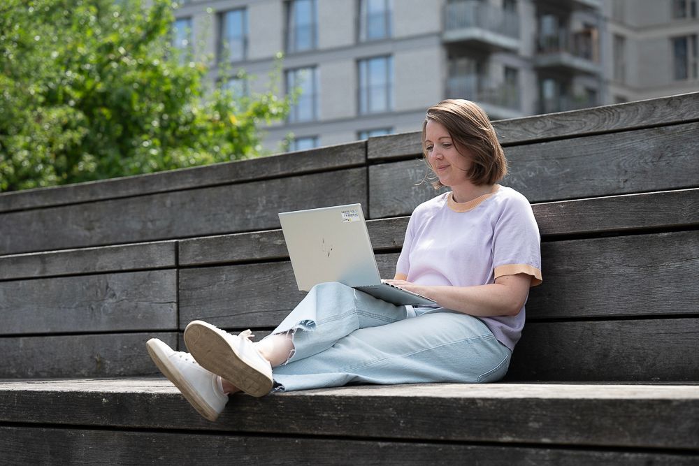 Eine Frau sitzt im Freien auf einer Holztreppe und benutzt einen Laptop. Sie trägt ein lavendelfarbenes Hemd, hellblaue Jeans und weiße Schuhe.