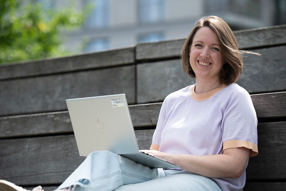 Eine Frau sitzt lächelnd draußen auf einer Holztreppe und arbeitet an ihrem Laptop.